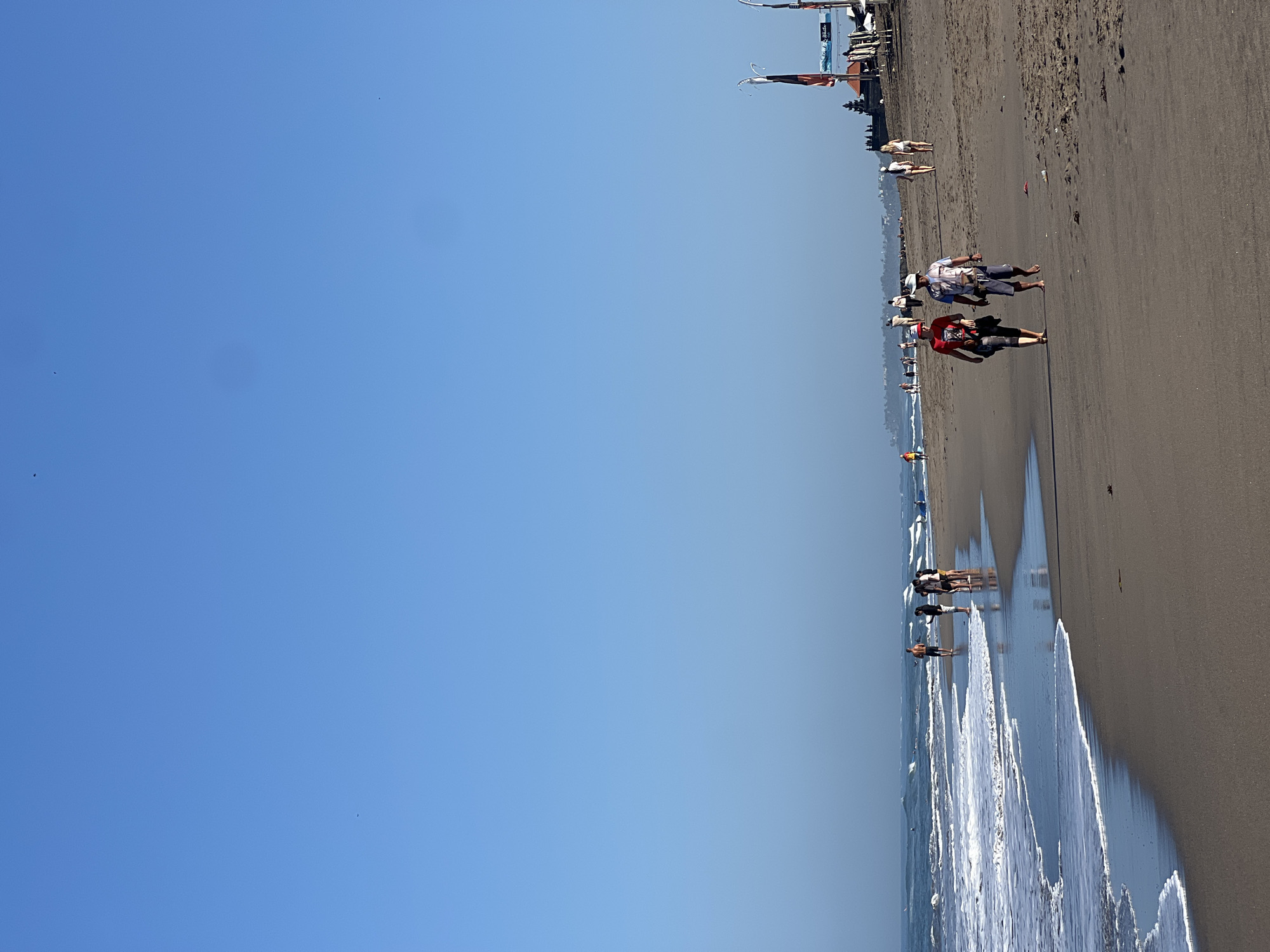 In North Kuta, Indonesia, a sandy beach stretches under a clear blue sky. Gentle waves lap the shore, creating white foam that recedes across the dark sand. Several people are visible walking along the beach. In the foreground, two individuals, one wearing a red shirt and the other a white and blue patterned shirt and a hat, walk side-by-side. Further down the beach, a group of four people stands near the water's edge, and other individuals are scattered further along the coastline. In the background, structures resembling buildings with flags and umbrellas suggest a populated area or resort. The overall scene depicts a tranquil beach environment with recreational activity.