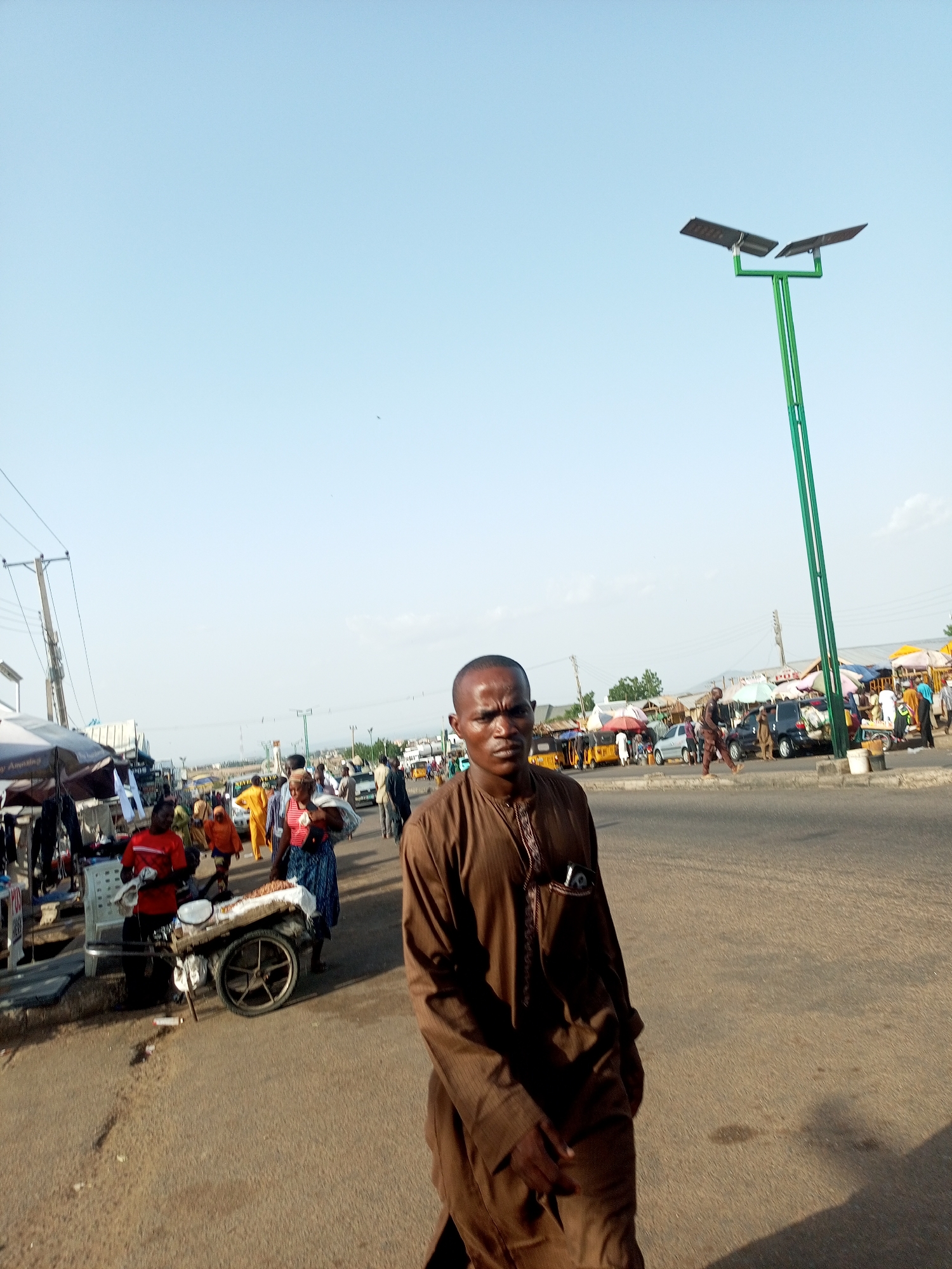 The image depicts a street scene in Gombe, Nigeria, likely a market area, under a bright, clear sky. A man in a brown traditional garment is walking towards the camera in the foreground, his gaze directed forward. To his left, a street scene unfolds with various individuals engaged in activities typical of a marketplace. Several people are seen walking, some carrying bags or goods. A cart laden with what appears to be food items is visible, with a vendor in a red shirt attending to it. Other stalls and vendors are set up further down the street, with umbrellas providing shade. Vehicles, including what looks like a commercial tricycle and cars, are also present, suggesting traffic and daily commerce. A tall, green solar-powered street light stands prominently on the right side of the frame. The ground is paved, with the texture of asphalt visible. The overall environment suggests a bustling, open-air market in an urban setting.