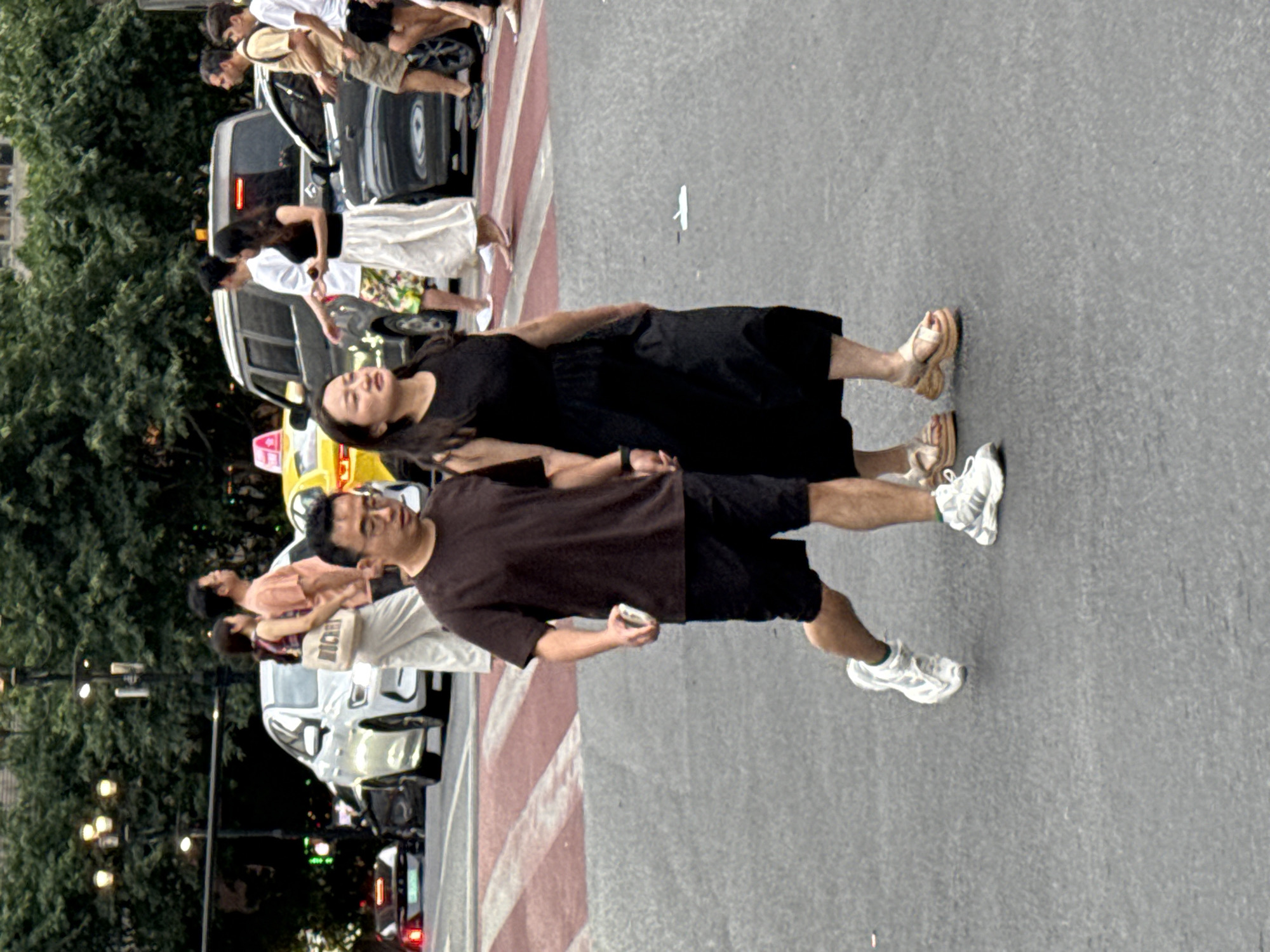 A man and a woman are walking hand-in-hand across a crosswalk in Yuzhong District, China. The man is wearing a dark t-shirt, black shorts, and white sneakers. The woman is wearing a black sleeveless dress and tan platform sandals. In the background, several cars, including a white SUV and a black sedan, are visible. Trees and streetlights line the street. The sky is overcast.