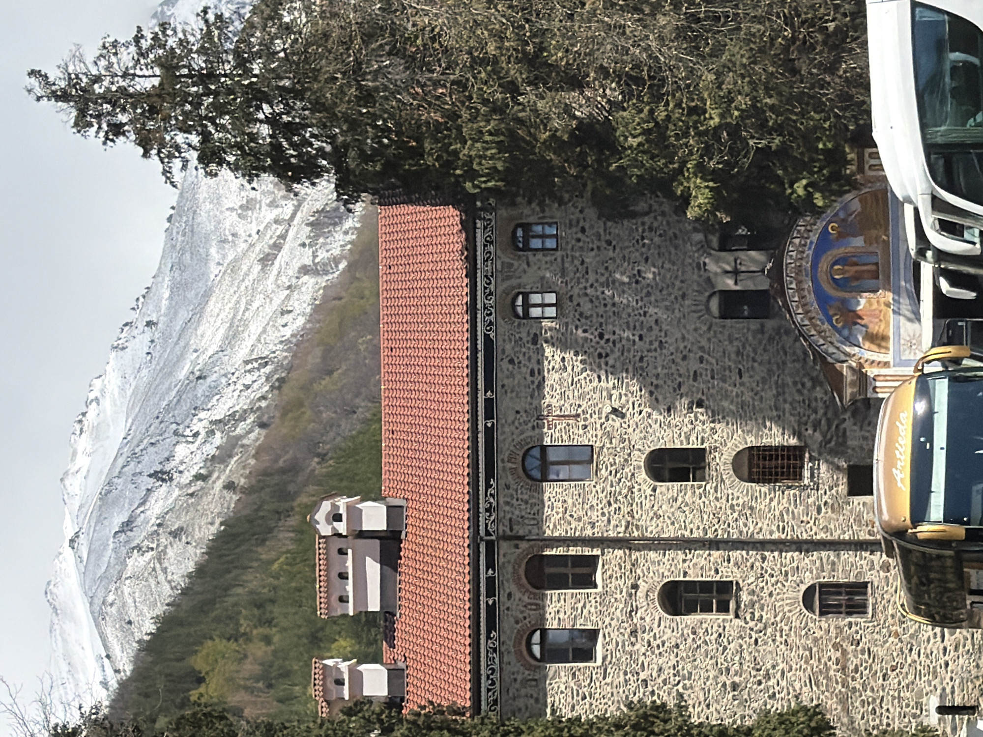 The image captures a view of Rila Monastery in Bulgaria, framed against a backdrop of a snow-covered mountain and a dense forest. The monastery building features stone walls with arched windows and a tiled roof. A section with what appears to be a small tower or addition is visible, also with the same tiled roof. A portion of a vehicle, possibly a bus or large van, is seen parked in front of the monastery. The surrounding environment includes trees and greenery. The overall impression is one of a historic building set within a striking natural landscape.