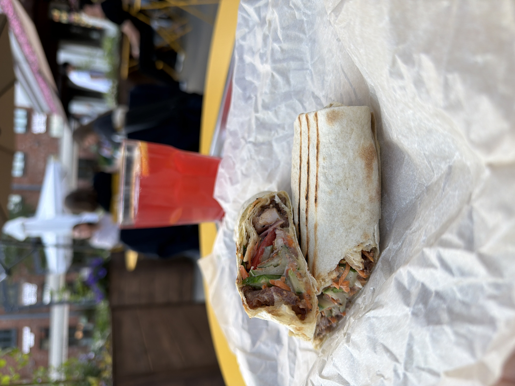 The image displays a close-up of a wrap, likely a shawarma or burrito, cut in half and resting on crumpled white paper. The filling is clearly visible, consisting of pieces of meat (possibly chicken or beef), shredded carrots, sliced cucumbers, and tomatoes, all mixed with a light-colored sauce. The tortilla shows distinct char marks from grilling. To the left, a glass containing a bright red beverage is partially visible, sitting on a vibrant yellow table.
The background is heavily blurred, indicating a shallow depth of field, and suggests an outdoor dining setting. Indistinct figures of people can be seen in the soft-focus background, some seated at tables and others standing. Vague elements of brick buildings and some green and purple foliage are also discernible.
The lighting indicates it is daytime, likely under bright or sunny conditions. The image is taken from a slightly tilted and elevated angle, focusing sharply on the food. The scene depicts an outdoor meal, possibly at a cafe, restaurant, or street food vendor. There is no visible text in the image.