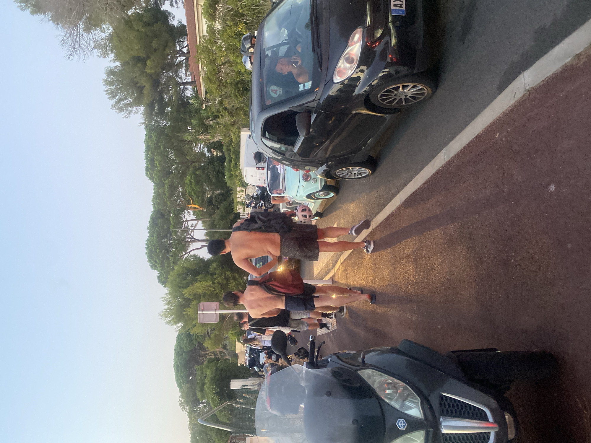 A sunny daytime street scene in Antibes, France, captures a line of people walking alongside a road. In the foreground, a black three-wheeled scooter is parked on the left. A group of individuals, primarily young men, are seen walking in single file on the sidewalk. One man in the foreground is wearing dark patterned shorts and carrying a backpack. Another man, behind him, is wearing navy blue shorts and also has a backpack. They appear to be walking away from the camera.
Further down the road, several vehicles are visible. A light blue retro-style car is in the middle lane, and a black Smart car is in the right lane. A person is visible driving the Smart car, looking out the window. Other parked vehicles and trees line the street, with buildings and more trees in the background, suggesting a suburban or coastal setting. The road surface appears to be asphalt, and the sidewalk has a reddish-brown textured surface. The overall atmosphere suggests a casual, relaxed environment, likely during warm weather given the attire of the people.