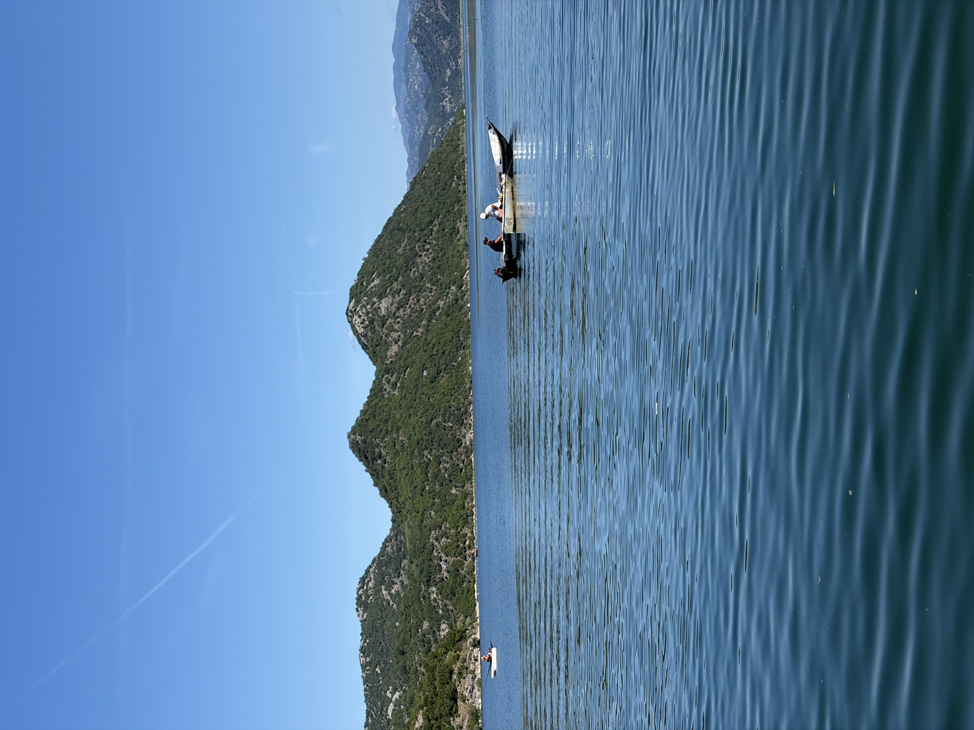 This image depicts a serene daytime scene over a large body of water, likely a lake, in the vicinity of Kruševica, Montenegro. The sky is a clear, bright blue with a single, prominent white contrail traversing it diagonally from the upper left. In the background, verdant, tree-covered hills rise from the water's edge, exhibiting rugged, rocky outcrops. Behind these immediate hills, further mountain ranges with lighter, hazy blue tones are visible on the horizon, suggesting distance.
The water, a deep blue-green, is calm with numerous small, parallel ripples stretching across its surface, reflecting the sunlight. Two boats are present on the water. To the left, a solo individual is seated in a small, white boat, resembling a kayak or canoe, oriented towards the right. This person appears to be wearing a light-colored hat and top.
Further to the right and closer to the center of the frame, a larger, traditional-style boat is stationary. This boat is light-colored, possibly white or grey, with an outboard motor mounted at its stern. Two individuals are inside this boat. The person on the left is wearing a dark cap and a dark shirt. The person on the right is wearing a white cap and a white shirt, positioned towards the front of the boat, seemingly interacting with something on board. The boat appears to be well-used, showing some wear and tear on its hull. The overall impression is one of peaceful outdoor activity, possibly fishing or recreation, amidst a natural landscape.