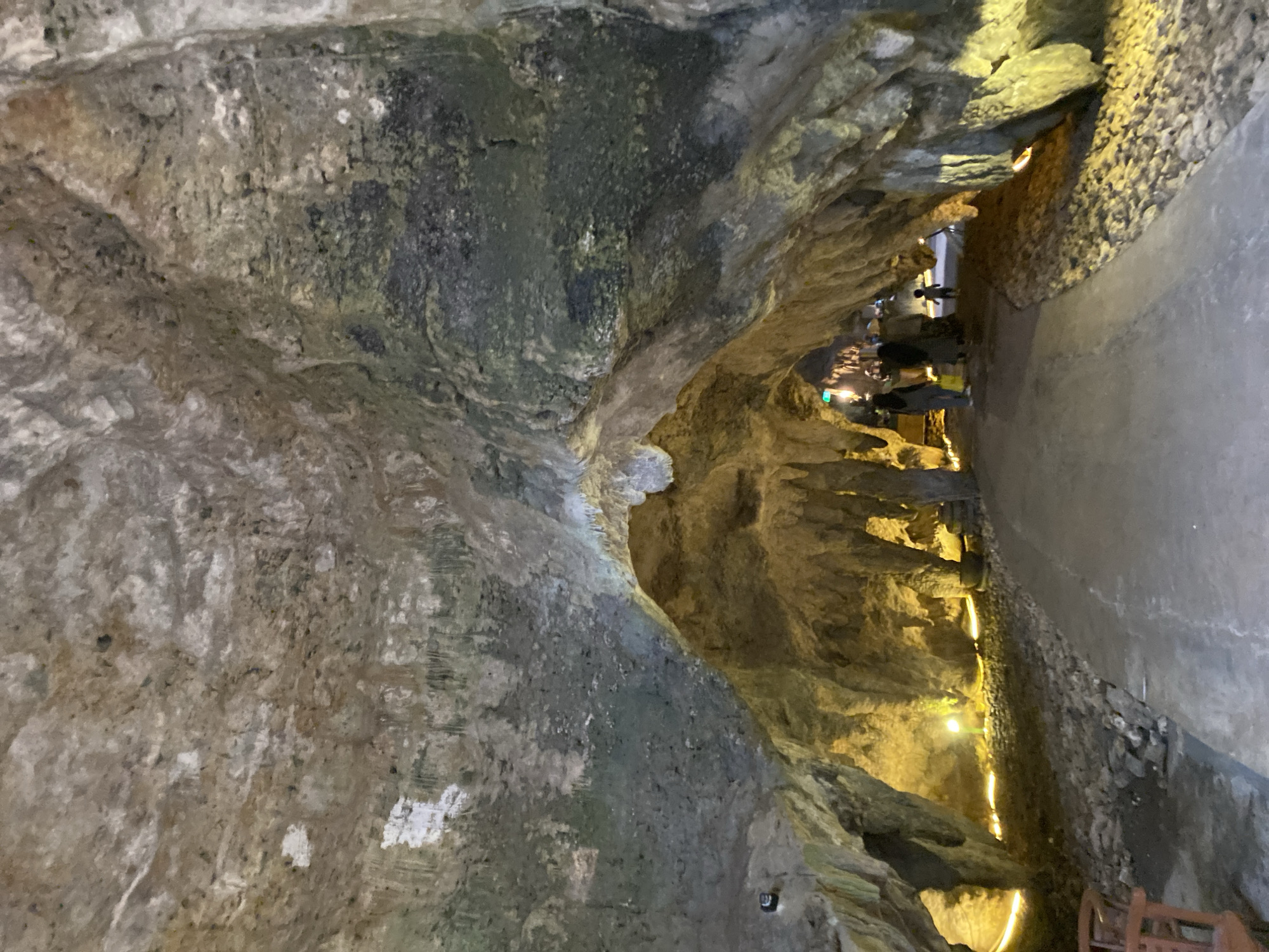 The image shows a view from inside a cave. The cave has a rough, textured ceiling and walls, with stalactites hanging from above and stalagmites rising from below in some areas. A pathway made of concrete winds through the cave. The pathway is lined with small stones on either side. Yellow lights are illuminating the pathway and the cave walls, creating a warm glow. In the background, a small group of people, including at least two adults and one child, are walking along the pathway. They are silhouetted against the light and their details are not discernible. The environment appears to be a tourist attraction within a cave.