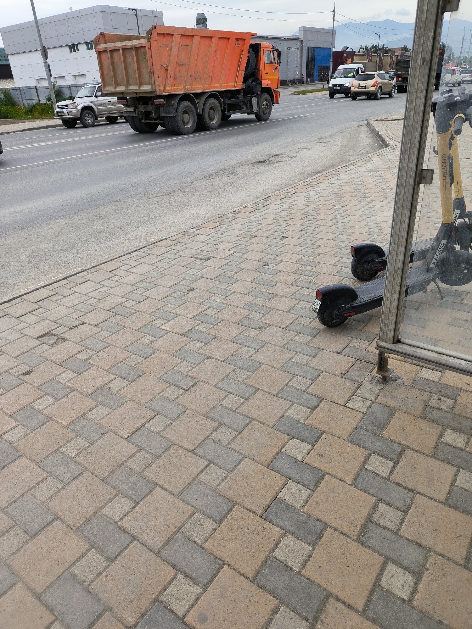 An orange dump truck is driving on a paved road in Yuzhno-Sakhalinsk, Russia. To the left of the truck, a silver SUV is also driving on the road. A sidewalk made of interlocking pavers is to the right of the road. Two electric scooters are parked on the sidewalk, next to a glass bus stop shelter. Buildings and distant mountains are visible in the background. The sky is overcast.