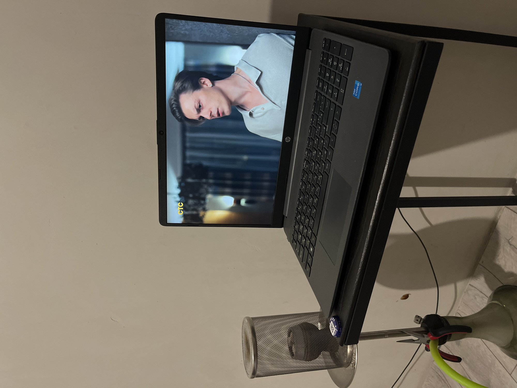 A laptop sits on a black table against a light beige wall. The laptop screen displays a scene from a movie or show featuring a light-skinned male with short dark hair, a silver necklace, and a light blue collared shirt. The laptop is partially open, revealing a black keyboard. A silver metal mesh cylinder is located to the lower left of the laptop, containing a dark object inside. Next to the cylinder is a hookah, consisting of a glass base, metal stem, and green hose. The image is taken indoors and appears to be in a residential setting. The exact location in the city of Verkhnyaya Pyshma, Russia, cannot be determined from the image.