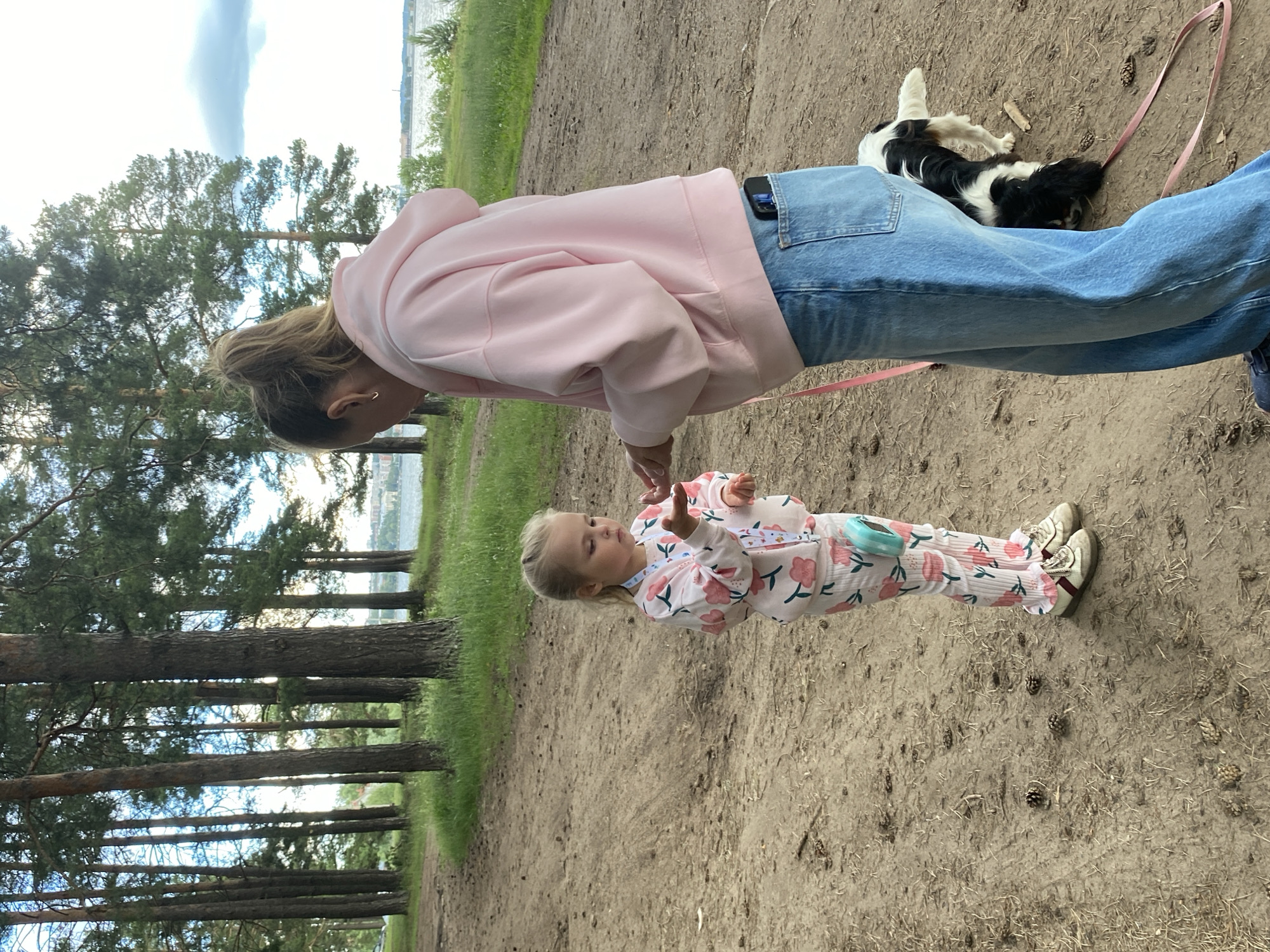 The image shows a woman and a young child walking outdoors in Votkinsk, Russia. The woman is wearing a pink hoodie and blue jeans, and is holding a pink leash. The child is wearing a floral-patterned outfit and white sneakers. A small black and white dog is lying on the ground near the woman's feet, attached to the leash.
They are in a wooded area with tall pine trees on one side and a grassy area leading to a body of water on the other. The ground is covered in dirt and pinecones, suggesting a natural, possibly park-like setting. The sky is overcast. The lighting suggests it is daytime, possibly late afternoon or early evening. The overall mood appears relaxed and casual, as the family enjoys an outdoor outing. There is no visible text in the image.