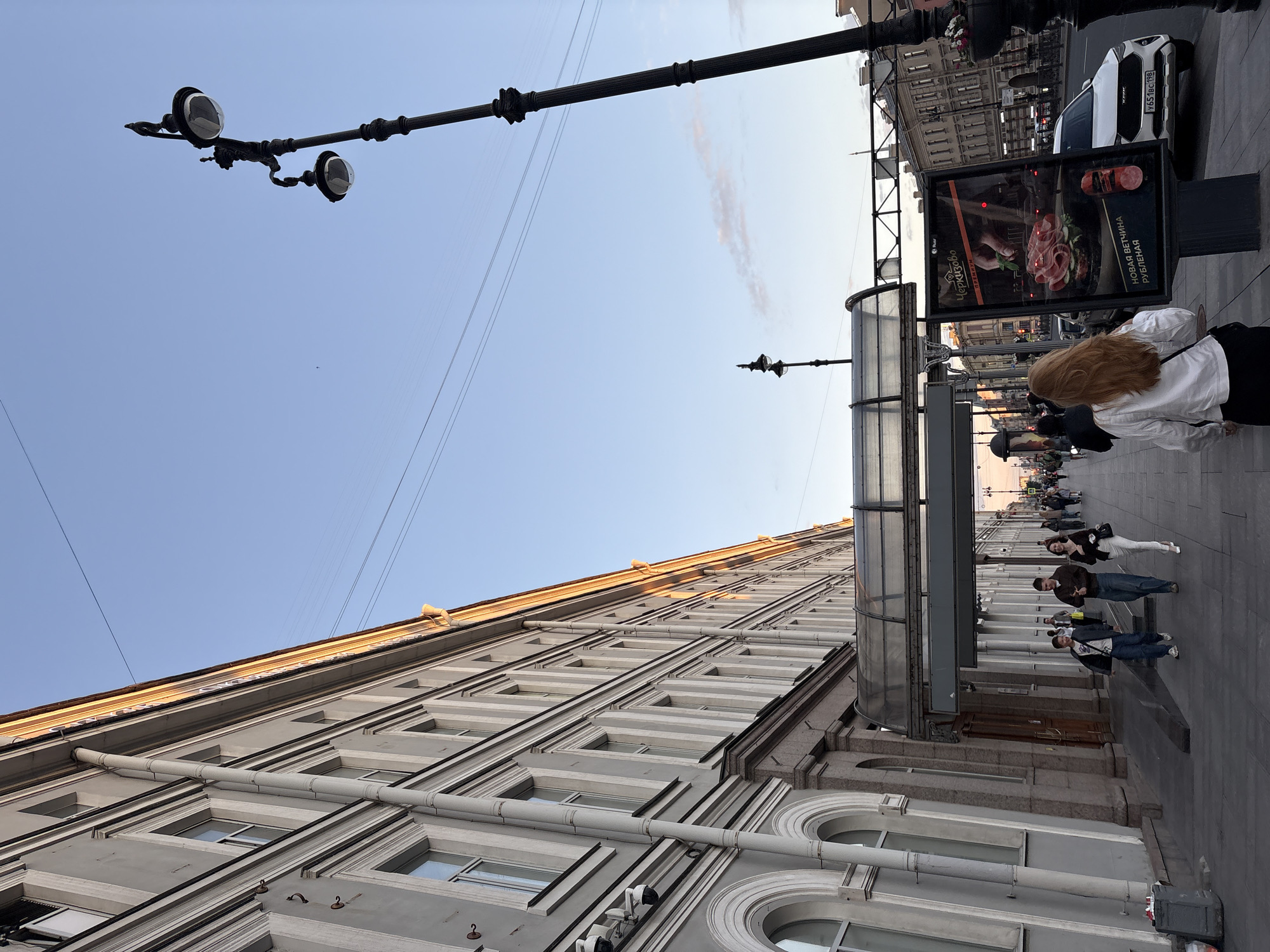 A medium-shot taken from a low angle shows a street scene in Saint Petersburg, Russia, during what appears to be late afternoon or early evening. The sky is a clear, pale blue with a few faint clouds. A tall, ornate black lamppost with two light fixtures stands prominently on the right side of the frame, silhouetted against the sky. The building on the left is a grand, multi-story structure with a light gray facade, featuring rows of windows with decorative moldings. Sunlight casts a warm glow on the upper portion of the building and the cornice.
In the foreground, a paved sidewalk is occupied by several pedestrians. Three people are walking towards the camera: a man in a blue jacket and jeans, a man in a brown jacket and dark pants, and a woman in a white shirt and dark pants, carrying a black bag. Further down the sidewalk, a larger group of people are walking away from the camera.
A clear glass and metal canopy extends from the building over the sidewalk. To the right of the canopy, a large advertising display is positioned. The display features an image of a sandwich with thinly sliced meat and an advertisement for "Cherkizovo" premium products, specifically "new diced bacon." A white car with a license plate that reads "Y651BC 198" is parked on the street behind the advertisement. The street beyond appears to be lined with more buildings and possibly other lampposts. The overall impression is of an urban street scene with a mix of architectural elements and pedestrian activity under a pleasant sky.