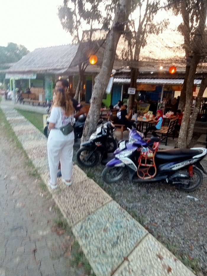 The image shows a scene in Jakenan, Indonesia, located at coordinates -6.758 latitude and 111.075 longitude. Three motorcycles are parked in front of a restaurant, one is purple and two are black. One person is standing in front of the motorcycles and two people are walking away. The restaurant is constructed with wood and has a thatched roof, shaded by a large tree. Tables and chairs are set up outside the restaurant, some people are sitting at the tables eating. There are several trees surrounding the restaurant.