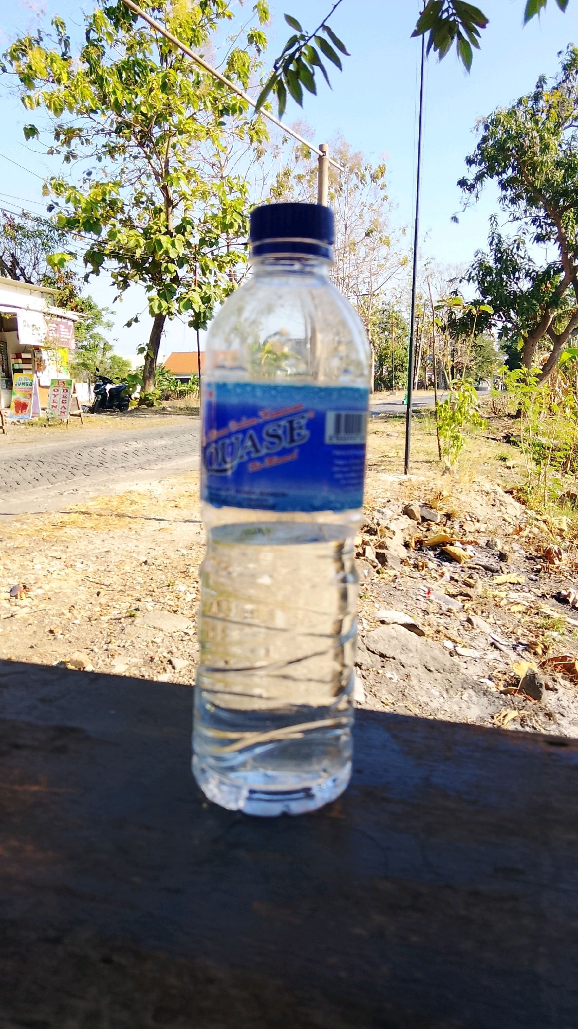 A partially filled plastic bottle of Aquase brand bottled water sits on a dark, wooden surface in the foreground. The bottle has a blue label with white text. The background shows a sunlit, outdoor scene with a paved road, dry, dusty ground, and trees with green leaves. A small building or stall with posters and a parked motorcycle is visible on the left side of the road. The sky is clear and blue. The image appears to have been taken in Dahanrejo, Indonesia, based on the context provided.