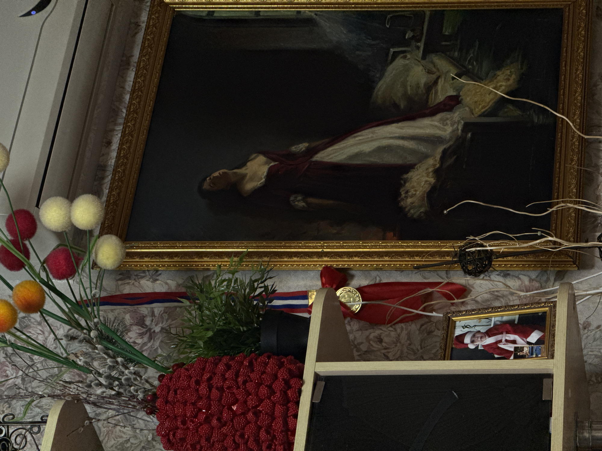 The image shows a framed painting in a gold frame, depicting a woman lying down. Below the painting, on a shelf, is an arrangement of artificial flowers and greenery, and a framed photograph of a baby dressed as Santa Claus. A gold medal is visible among the artificial flowers. The wall behind the objects has a floral pattern. The location is unknown.
