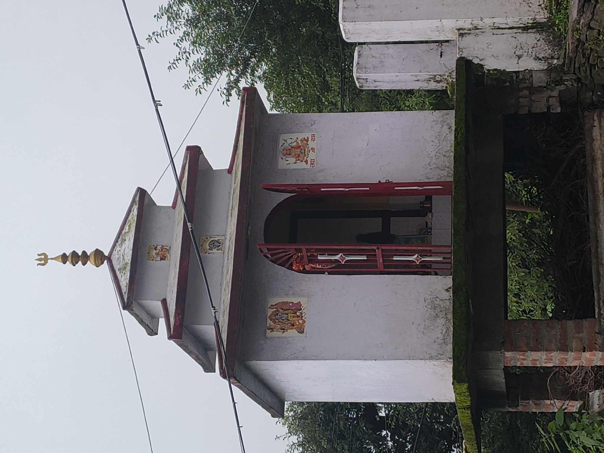 A small, two-tiered shrine building, painted white with red trim, stands on a concrete platform supported by a brick structure. A golden trident finial tops the shrine. Red metal doors, partially open, reveal a dark interior. The shrine is decorated with religious images on its exterior walls, including depictions of Hindu deities and text. The structure is situated outdoors, with lush green trees and foliage surrounding it. Power lines are visible overhead. The shrine appears to be located in a rural or semi-rural setting, possibly in the town of Haini, India, given the architectural style and the presence of religious iconography common in the region. The overall scene suggests a place of worship in a natural environment.