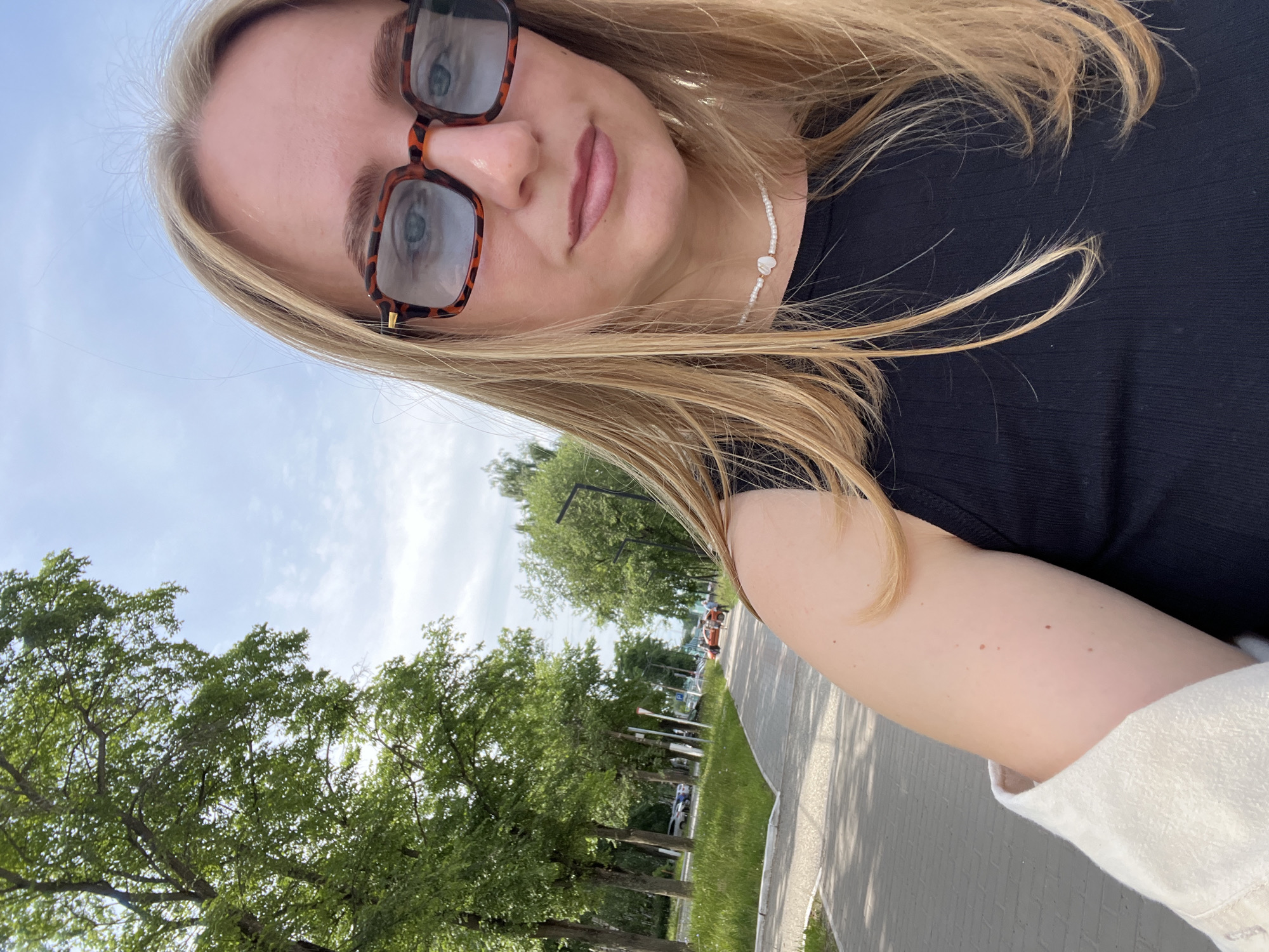 A young woman with blonde hair and tortoise-shell glasses takes a selfie outdoors. She is wearing a black tank top and a light-colored jacket draped over her shoulder. A beaded necklace with a pearl pendant adorns her neck. Behind her, a paved walkway lined with trees stretches into the distance, with a street and parked cars visible on the left. The sky is partly cloudy. The scene appears to be a public park or promenade in Chaikovsky, Russia.