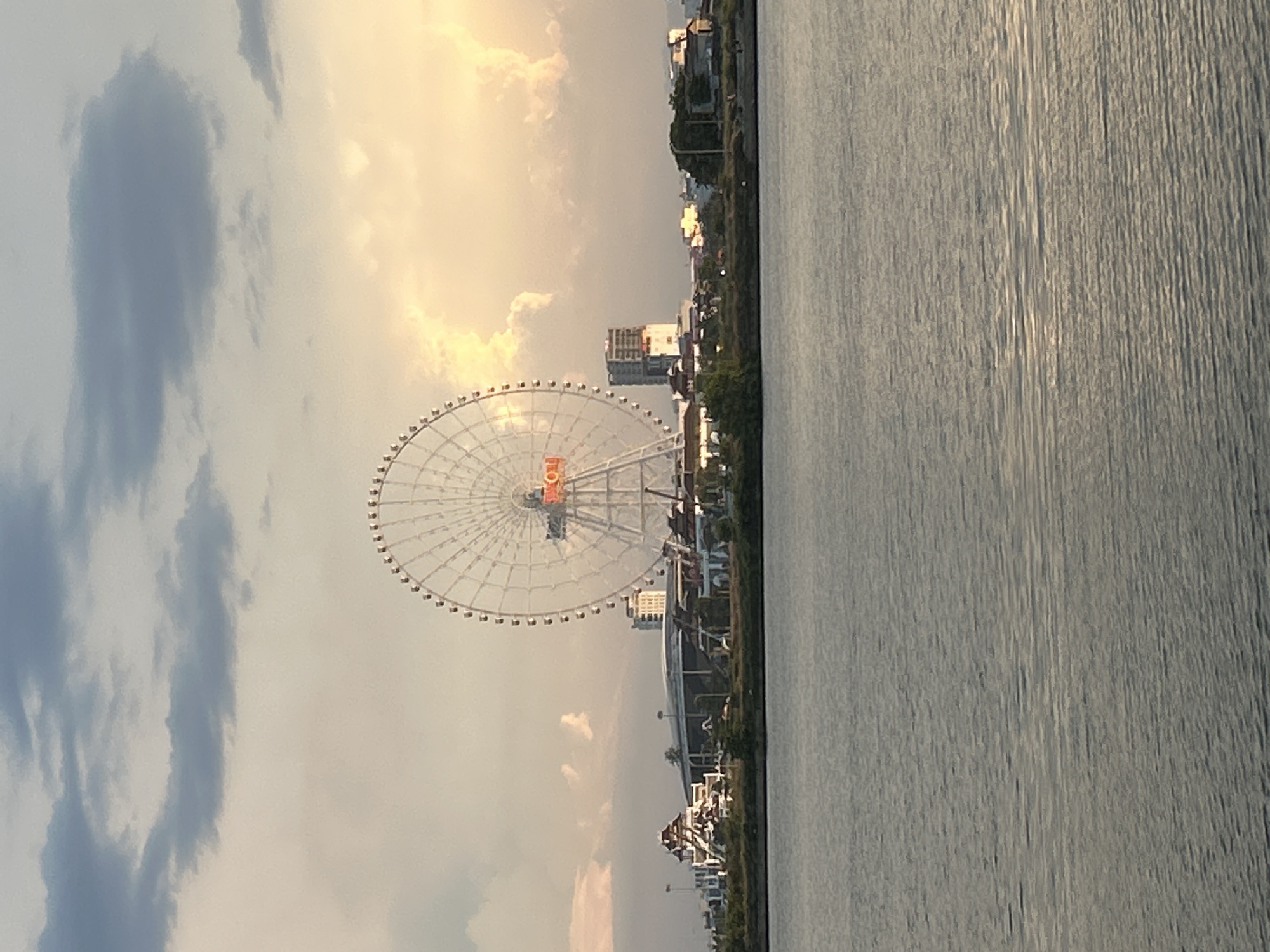 The image captures a scene along the Han River in Da Nang, Vietnam. Dominating the view is a large Ferris wheel, illuminated with orange and red lights that spell out "SUN". The Ferris wheel stands on the bank of the river, with a cityscape visible behind it, including modern buildings and what appears to be an amusement park or entertainment complex. The sky above is a dramatic mix of soft, warm-toned clouds tinged with sunset light and darker, brooding clouds. The foreground is occupied by the calm, rippling surface of the Han River, reflecting the soft light of the sky. The overall atmosphere is serene and picturesque, suggesting a late afternoon or early evening setting.