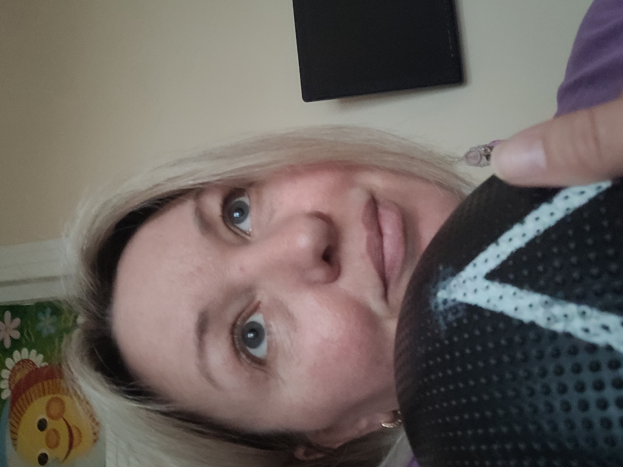 A selfie features a woman with light blonde hair and light complexion holding a black ball with a white triangle design. Her right index finger rests on the ball's surface. Her eyes are blue-gray. To the left of the woman, part of a child's wall decoration is visible, with a drawing of a figure wearing a red and yellow hat. Behind her, slightly above, there is a black rectangular object mounted on the wall. The background is light beige. There is no indication of the city of Izhevsk, Russia in this image.