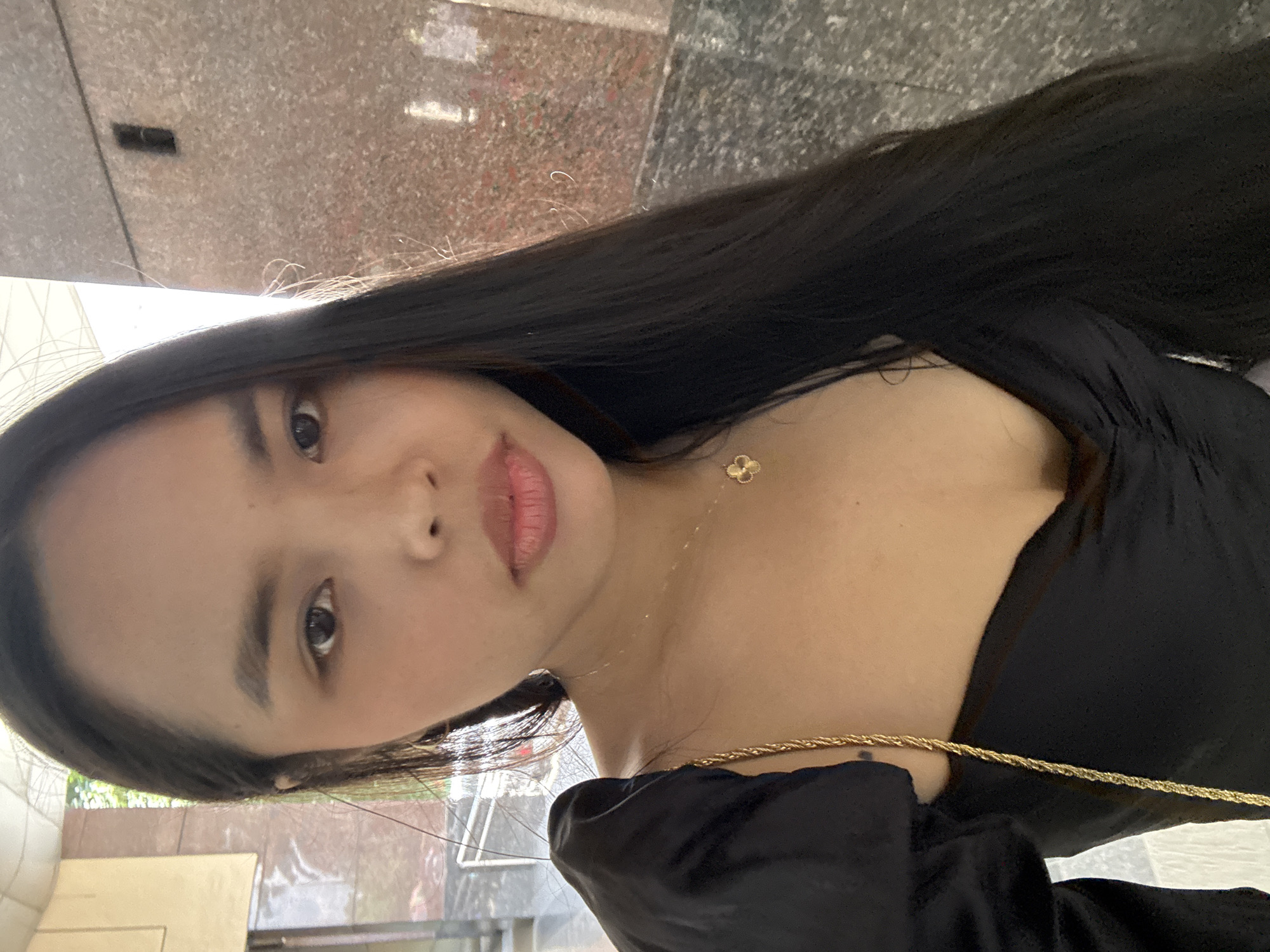 A young woman with long, dark, straight hair is captured in a close-up, chest-up shot, looking directly at the camera. She has a neutral to slightly smiling expression with her lips lightly parted, revealing a soft pink lip color. Her skin tone is warm, and her dark eyes are prominent. She is wearing a black top, possibly a dress or blouse, with a draped or ruched detail around the neckline.
Around her neck, she wears a delicate gold-colored chain necklace with a small, four-leaf clover-shaped pendant. Another thicker, gold-colored braided chain or strap is visible near her right shoulder, possibly part of a bag or accessory.
The background is softly blurred but appears to be an indoor or semi-outdoor urban setting, characterized by textured, possibly stone or marble walls, and reflective surfaces that suggest a building interior or facade. Hints of light and indistinct architectural elements are visible in the background, suggesting a daytime setting. The image provides no visible text. The location context for this scene is Davao City, Philippines.