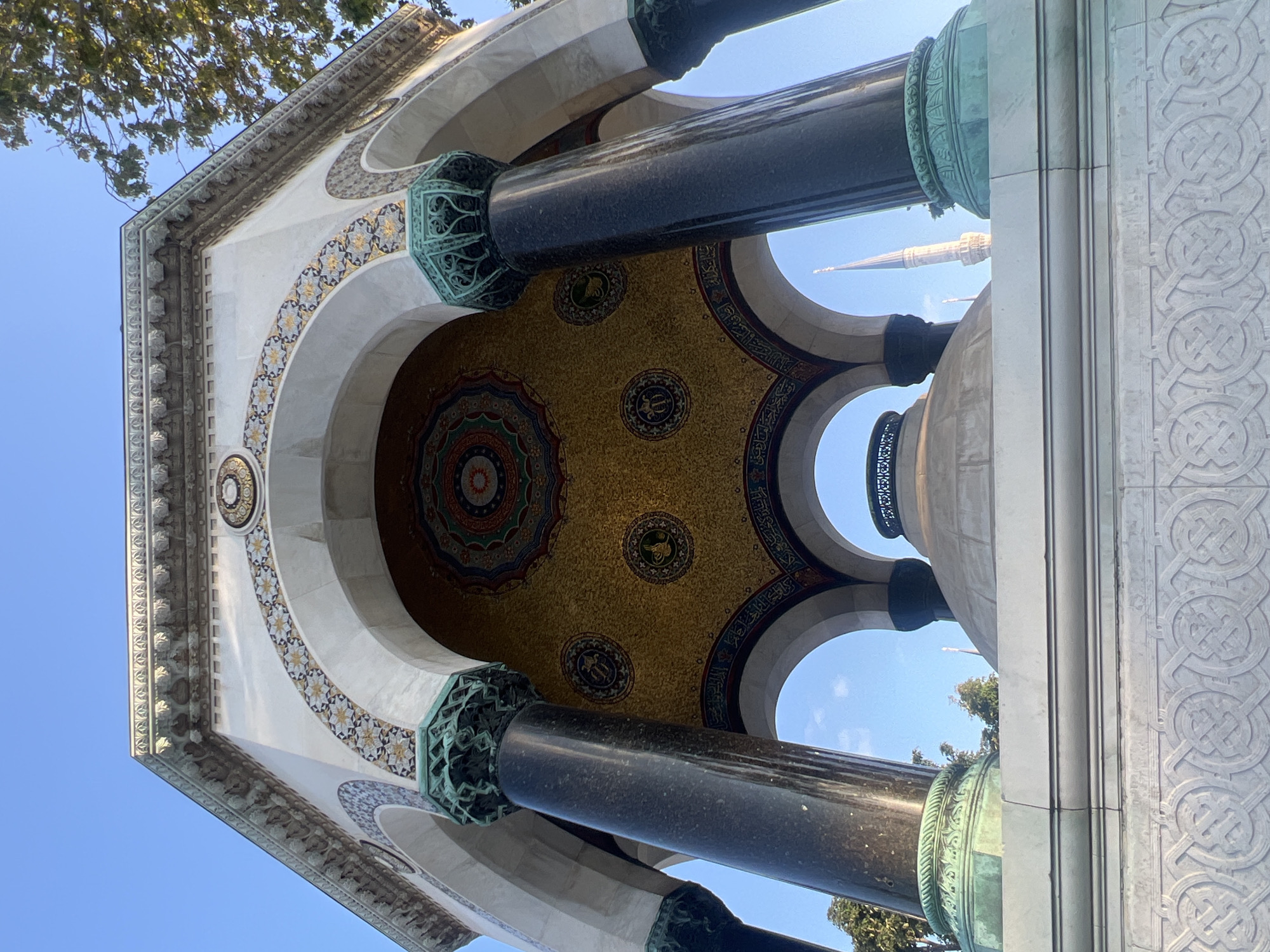 The image shows the German Fountain in Sultanahmet Square, Istanbul, Turkey. The fountain is a hexagonal structure with a domed roof. It is made of marble and decorated with mosaics and calligraphy. The fountain has four arches, through which a minaret and trees are visible in the background. The sky is clear and blue. There are no people in the image.