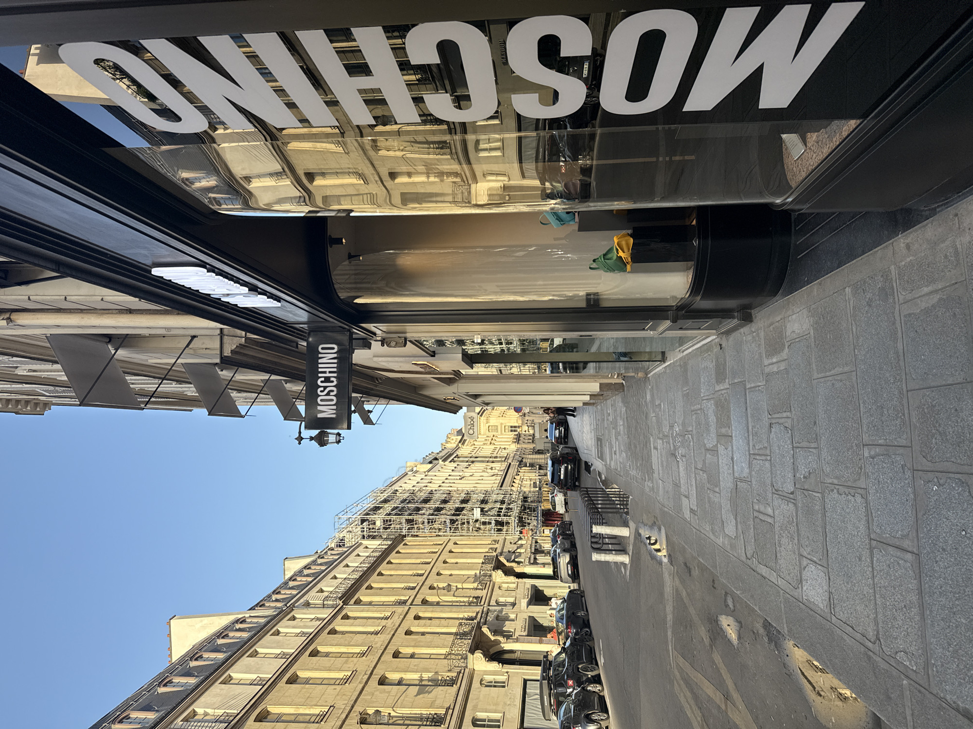 The image depicts a street in Paris, France, with a Moschino store on the left. The store's facade features the brand's name in large white letters, and its window displays are visible. The street is lined with traditional Parisian buildings with multiple windows and ornate details. Scaffolding is present on one of the buildings, suggesting ongoing construction or renovation. Cars are parked along the street, and pedestrians can be seen in the distance. The sky is clear and blue. The pavement is made of stone blocks.