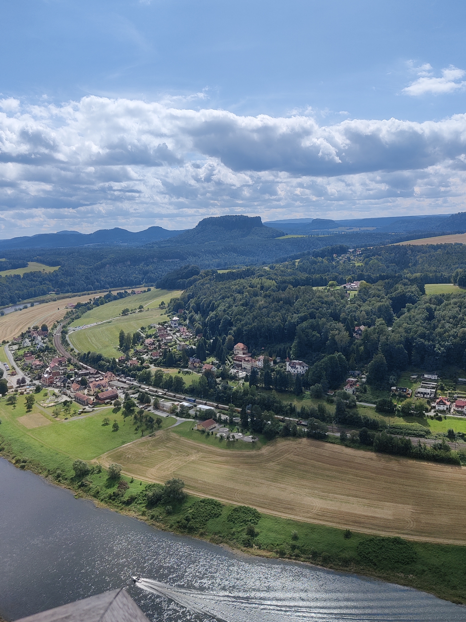 This is an aerial photograph of the Elbe Sandstone Mountains region, near the town of Lohmen, Germany. In the foreground, a dark river flows from the left to the right, with a small white boat creating a wake on its surface. The riverbank is lined with green grass and trees.
In the middle ground, a village with red-tiled roofs is nestled along a curved railway track. The track is occupied by a long train of blue carriages. Fields of dry, harvested crops are visible behind the village, transitioning into dense green forests that cover the rolling hills. Several houses with white facades and red roofs are scattered throughout the treeline.
In the background, a series of layered mountain ranges stretch towards the horizon under a bright blue sky filled with scattered white clouds. The overall impression is a picturesque rural landscape with a clear division between the river, the village, and the natural surroundings.