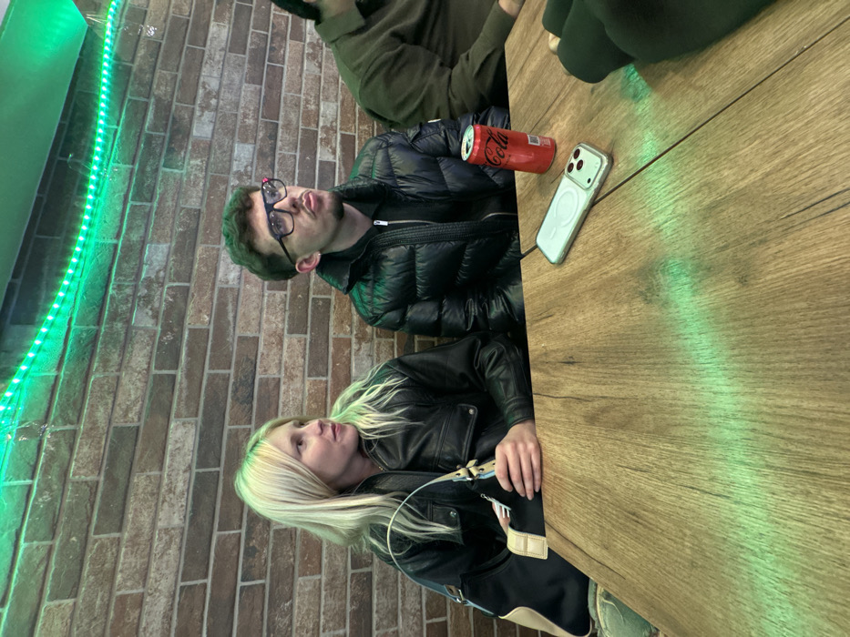 Three people are seated at a wooden table next to a brick wall illuminated with green LED lights. A man wearing glasses and a black puffer jacket sits next to a can of Coca-Cola and an iPhone. A woman with blonde hair, dressed in a black leather jacket, is also at the table. A third person wearing a green top is partially visible.