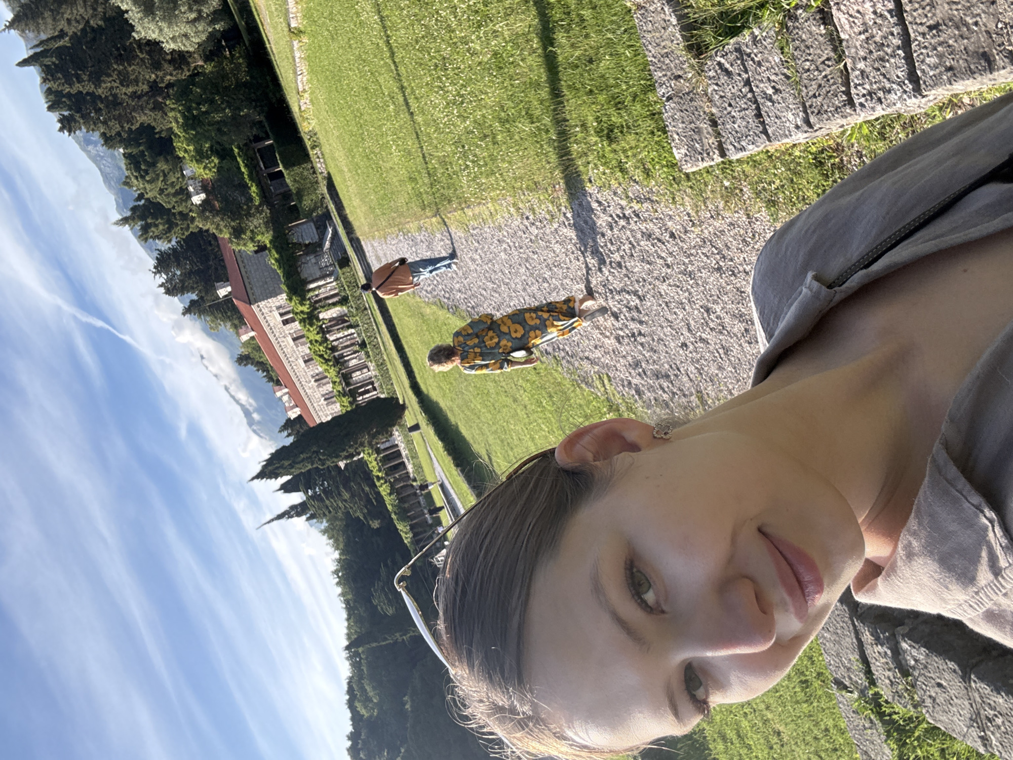 A selfie is taken outside the Villa Miločer in Sveti Stefan, Montenegro. The photographer is a woman with light skin and brown hair. Behind her, two people stroll along a gravel path. One wears a floral-patterned jacket, and the other a brown jacket and blue jeans. A manicured green lawn extends to a stone building with red roofing and vegetation growing on its walls. The sky is blue with streaks of clouds.