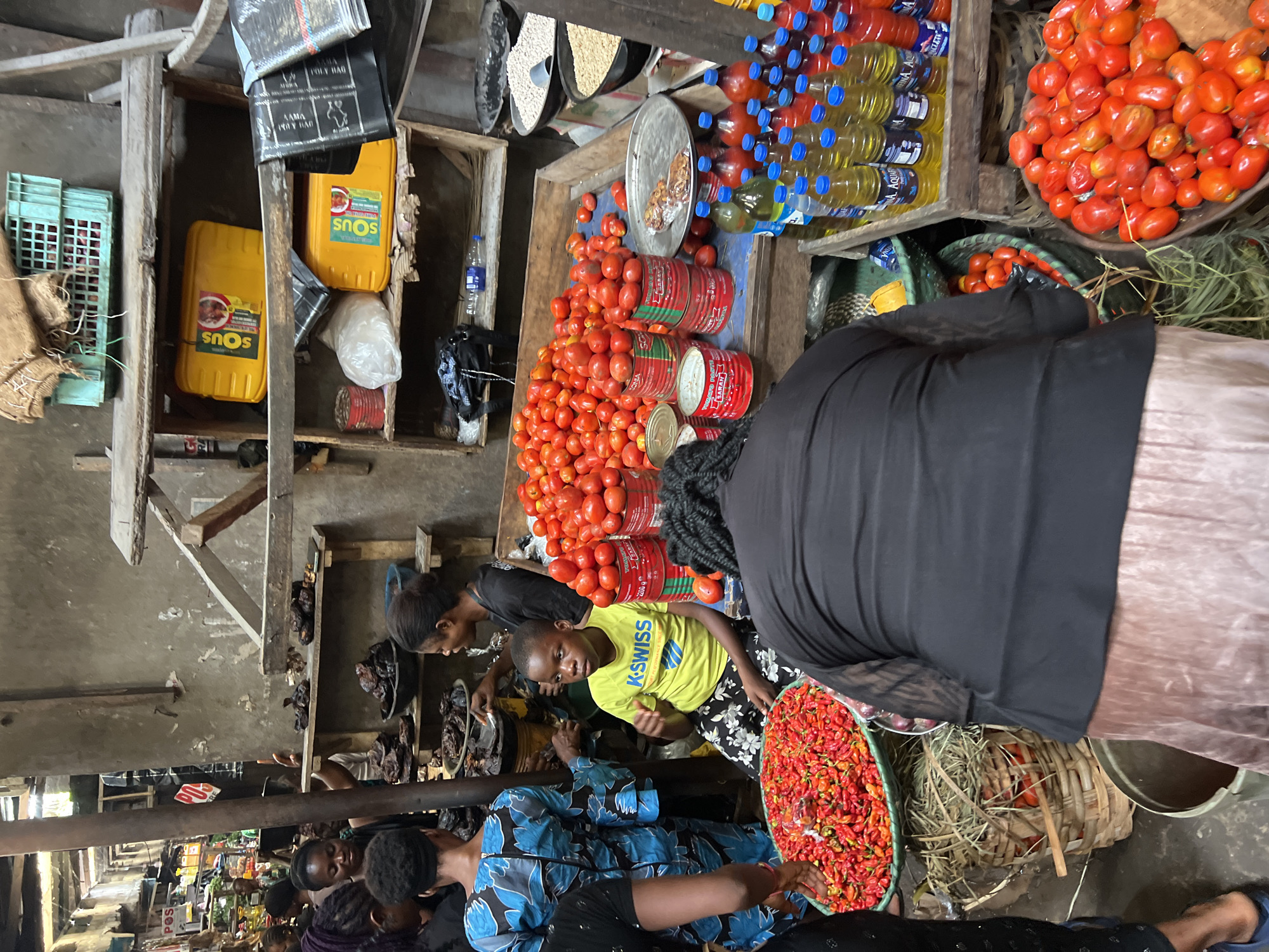 The image depicts a bustling outdoor market scene in Nkpor, Nigeria. In the foreground, a young boy wearing a bright yellow t-shirt with "K-SWISS" printed on it is seated in front of a large basket overflowing with red chili peppers. To his right, a woman with braided hair is obscured from view, but her back is turned to the camera. Behind the boy, a mound of ripe red tomatoes is displayed. Further back, several other individuals are visible, engaged in market activities. Some are examining produce, while others appear to be vendors. Shelves in the background hold various goods, including yellow containers of cooking oil, bags of rice, and dried goods. The market environment is characterized by wooden stalls and overhead structures, suggesting an informal and lively marketplace. The overall impression is one of a vibrant commercial hub in Nkpor, Nigeria.