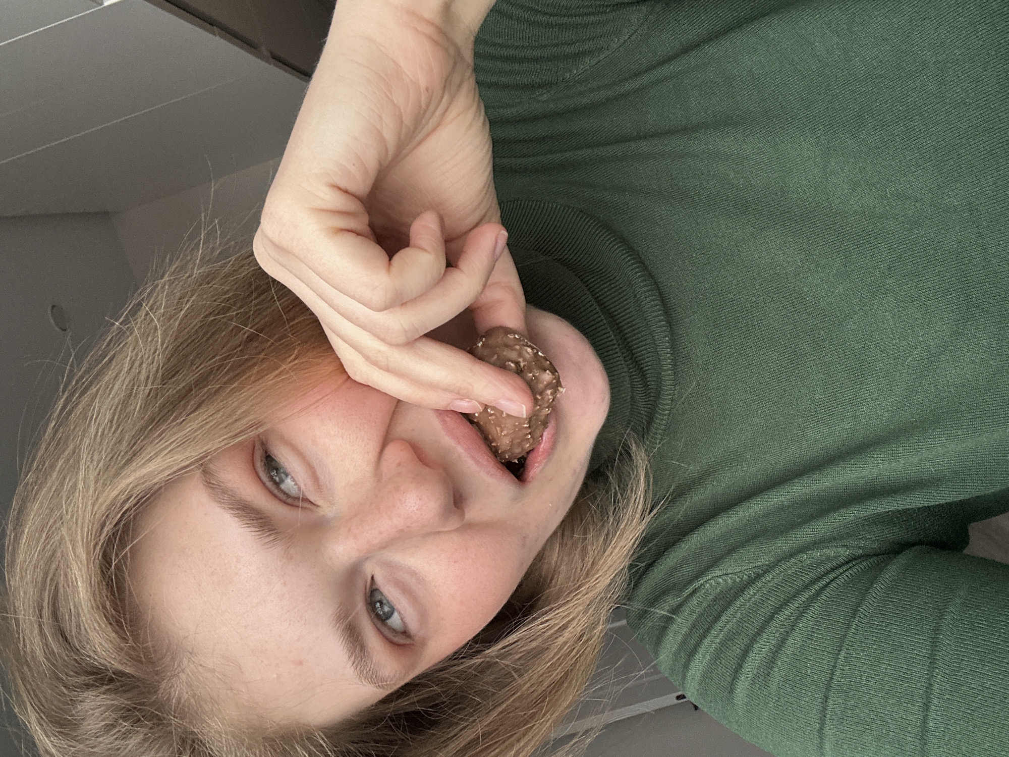A young, fair-skinned person with blonde hair and blue eyes is holding a chocolate candy in their mouth. The person is wearing a green turtleneck sweater. The background is a light gray color, possibly a ceiling. The picture is taken from a low angle, looking up at the person. The location is not identifiable as Kazan, Russia, from the given image.