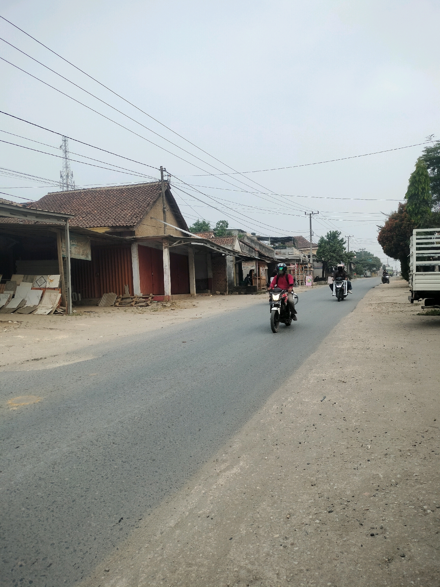 The image depicts a street scene in the city of Wayakroy, Indonesia. A paved road runs through the frame, with buildings lining the left side and a dirt shoulder on the right. Several motorcycles are present. One motorcycle in the foreground, driven by a person wearing a red shirt and a helmet, is moving away from the camera. Further down the road, two more motorcycles are visible, also moving away from the camera. On the right side of the road, a white truck with a flatbed is parked. The buildings on the left appear to be shops or houses, with some displaying goods outside. Power lines and poles are visible overhead. The sky is overcast.
