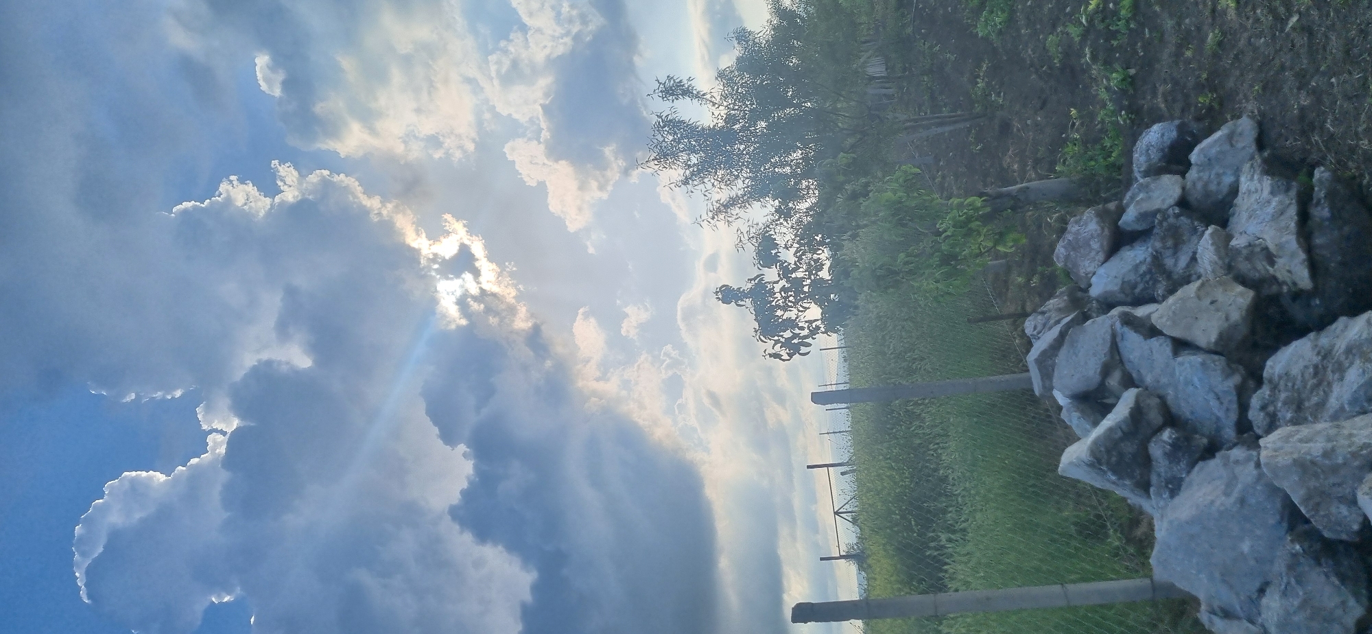 The image captures a rural landscape with a vibrant sky. A pile of gray rocks is situated in the lower right corner of the frame. A wire fence runs horizontally across the midground, supported by metal posts. Beyond the fence, lush green vegetation covers the ground. A tree with sparse leaves stands to the right of the frame. The sky is filled with dramatic, cumulus clouds, partially obscuring the sun, which casts rays of light. The location appears to be a rural area.