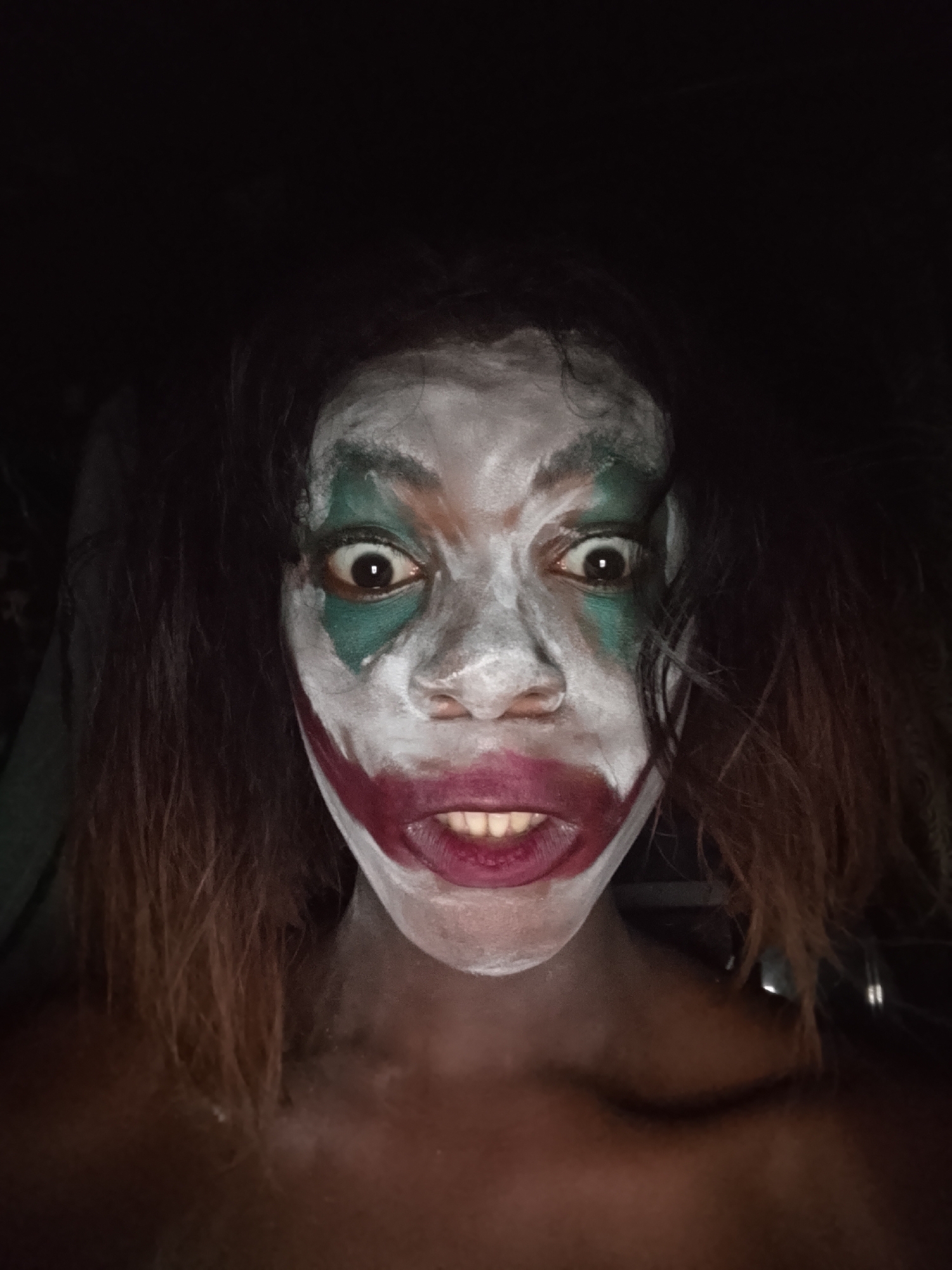 A close-up portrait features a person with dark skin and dark, reddish-brown hair, adorned with intricate clown-like face paint reminiscent of the Joker character. The face is painted predominantly white, with vibrant green accents around the eyes, extending outwards and downwards from the inner corners. A bold, dark red or burgundy color creates an exaggerated, wide smile, stretching from the corners of the mouth across the cheeks.
The person is looking directly into the camera with wide, intense eyes, one of which appears slightly larger and more startled. Their mouth is open, revealing teeth, contributing to an unsettling and manic expression. The hair, long and dark, frames the face and appears somewhat dishevelled.
The setting is dimly lit, with the background almost entirely dark, suggesting the photograph was taken in a dark room or at night. The poor lighting further emphasizes the stark contrast of the white and colored makeup against the dark surroundings and the person's skin tone. The overall mood conveyed is one of intensity, surprise, and a touch of the macabre, typical of horror or Halloween aesthetics. No visible text is present in the image.