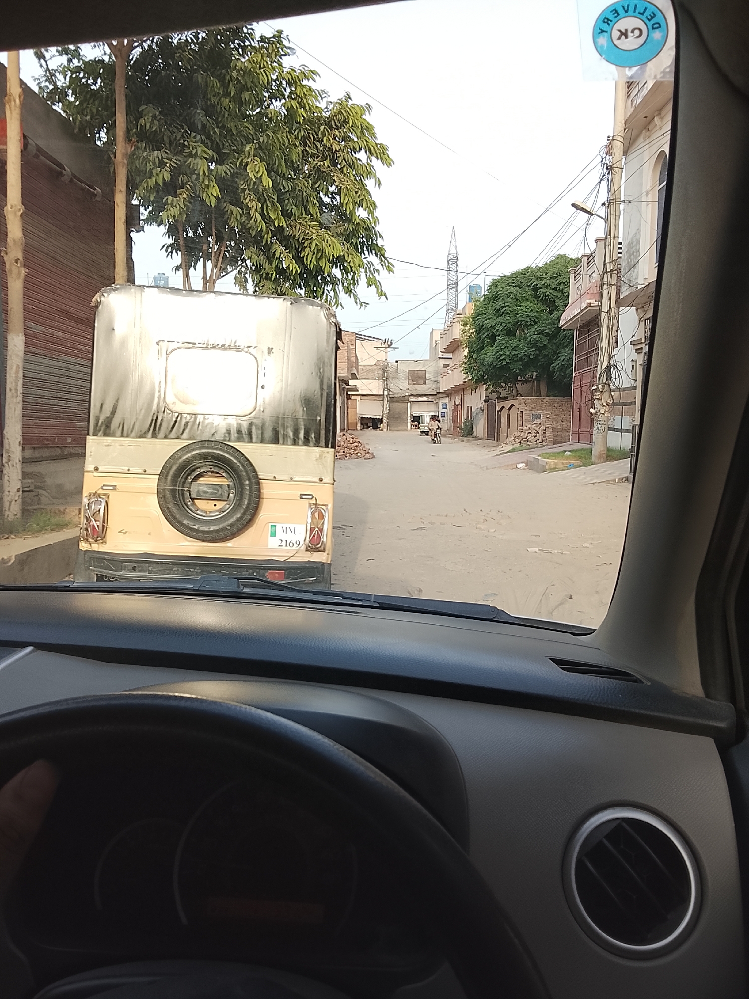 The image shows a view from inside a car, looking out onto a street in Nasheman Colony, Pakistan. The main subject is a three-wheeled vehicle (rickshaw or auto-rickshaw) directly in front of the car. The rickshaw is beige with a covered passenger area and a spare tire mounted on the back. It has a license plate with the designation "MNL 2169". The street appears unpaved and dusty. Buildings are visible on both sides of the street, with a mix of residential and commercial structures. Some buildings have brick or concrete exteriors. Utility poles with wires are also present. In the distance, a tall lattice tower or communication tower is visible. In the far distance, a person is on a motorcycle.