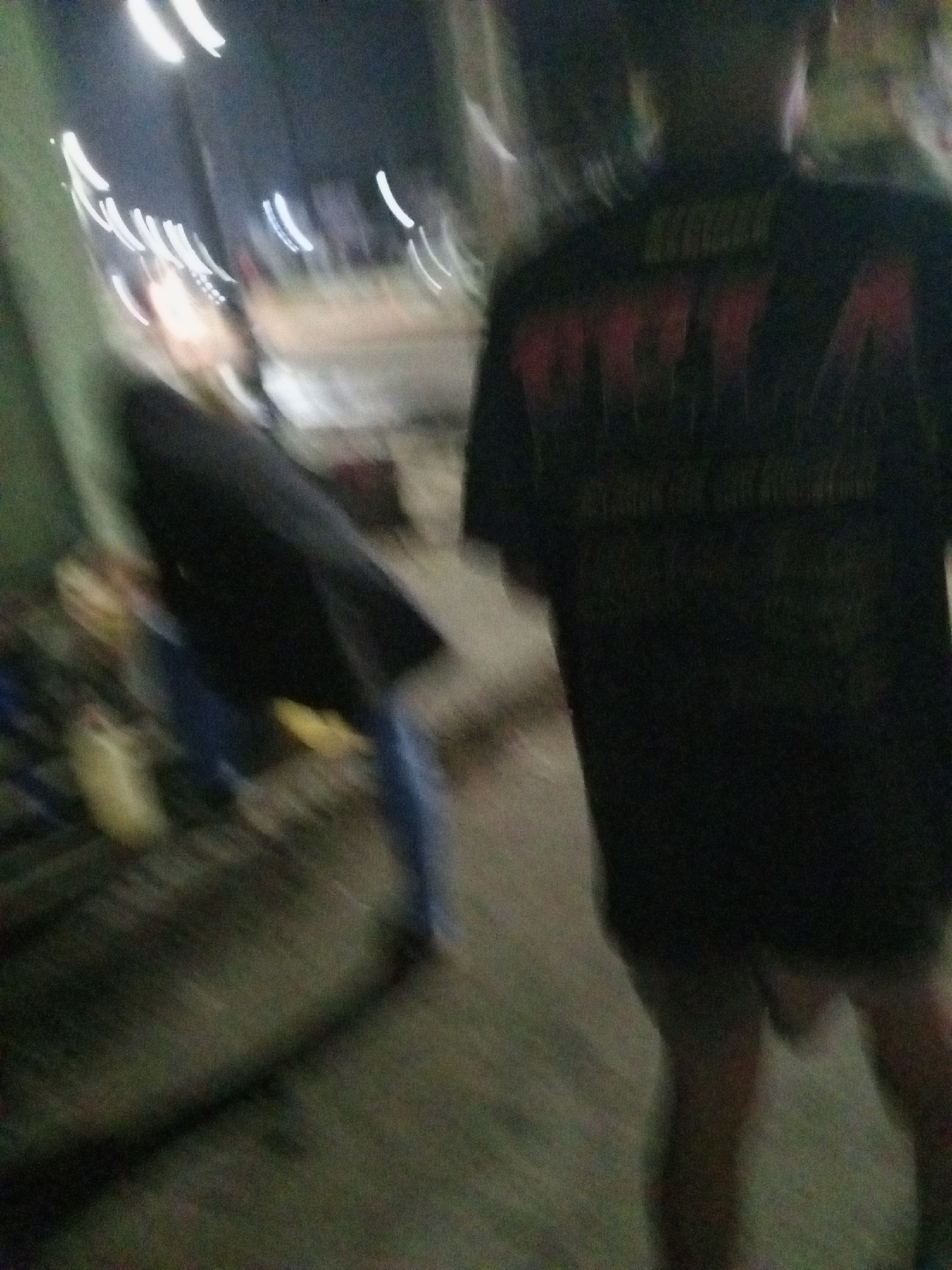 The image depicts a blurry scene taken outdoors at night, possibly in Ado, Nigeria.
A person wearing a dark t-shirt with a possible graphic print on the back is standing facing away from the camera. They are wearing shorts.
To the left, there is a blurred figure wearing a dark jacket and jeans. In the background, there appear to be lights, possibly from street lamps or buildings. The ground appears to be a sidewalk or paved area. There is foliage on the left. The image is affected by motion blur, obscuring details and making the scene difficult to discern.