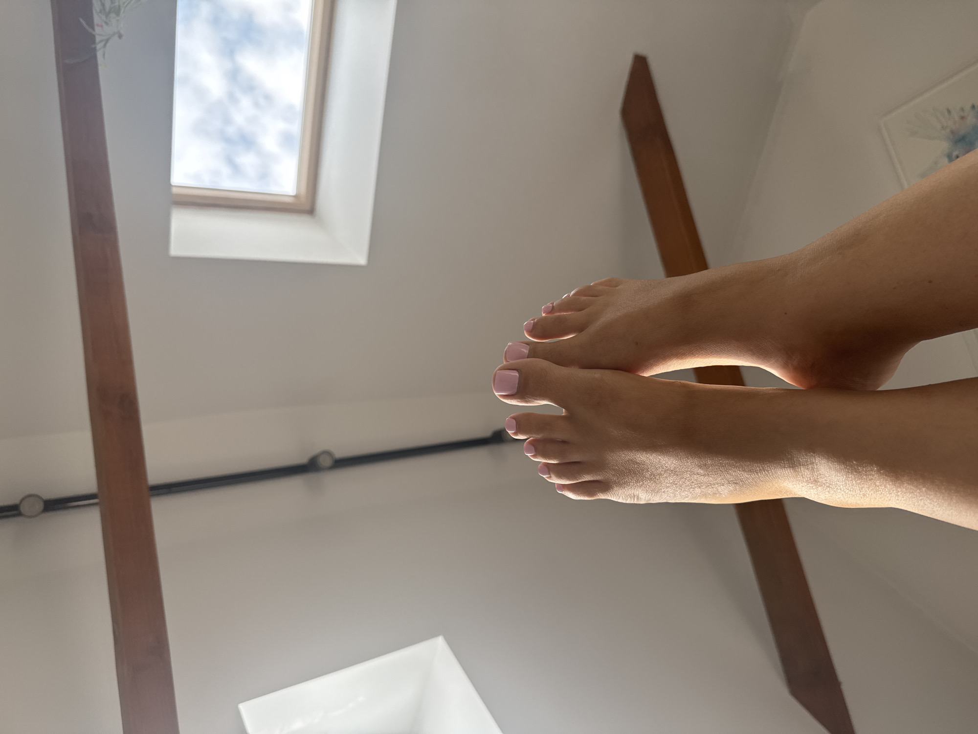 The image shows a close-up, upward-facing view of a person's feet and lower legs. The feet are positioned in the foreground, with toes pointed upwards. The toenails are painted a pale pink color. The skin of the legs and feet appears to be tanned.
The background is an interior ceiling with exposed wooden beams and what appear to be skylights. One skylight shows a blue sky with white clouds. There is also a black conduit running along the ceiling.
The context of the image being in Katowice, Poland, is not visually apparent from the image itself. The scene depicts a domestic or indoor setting. The focus is on the subject's feet and legs, with the architectural elements of the ceiling and windows forming the backdrop.