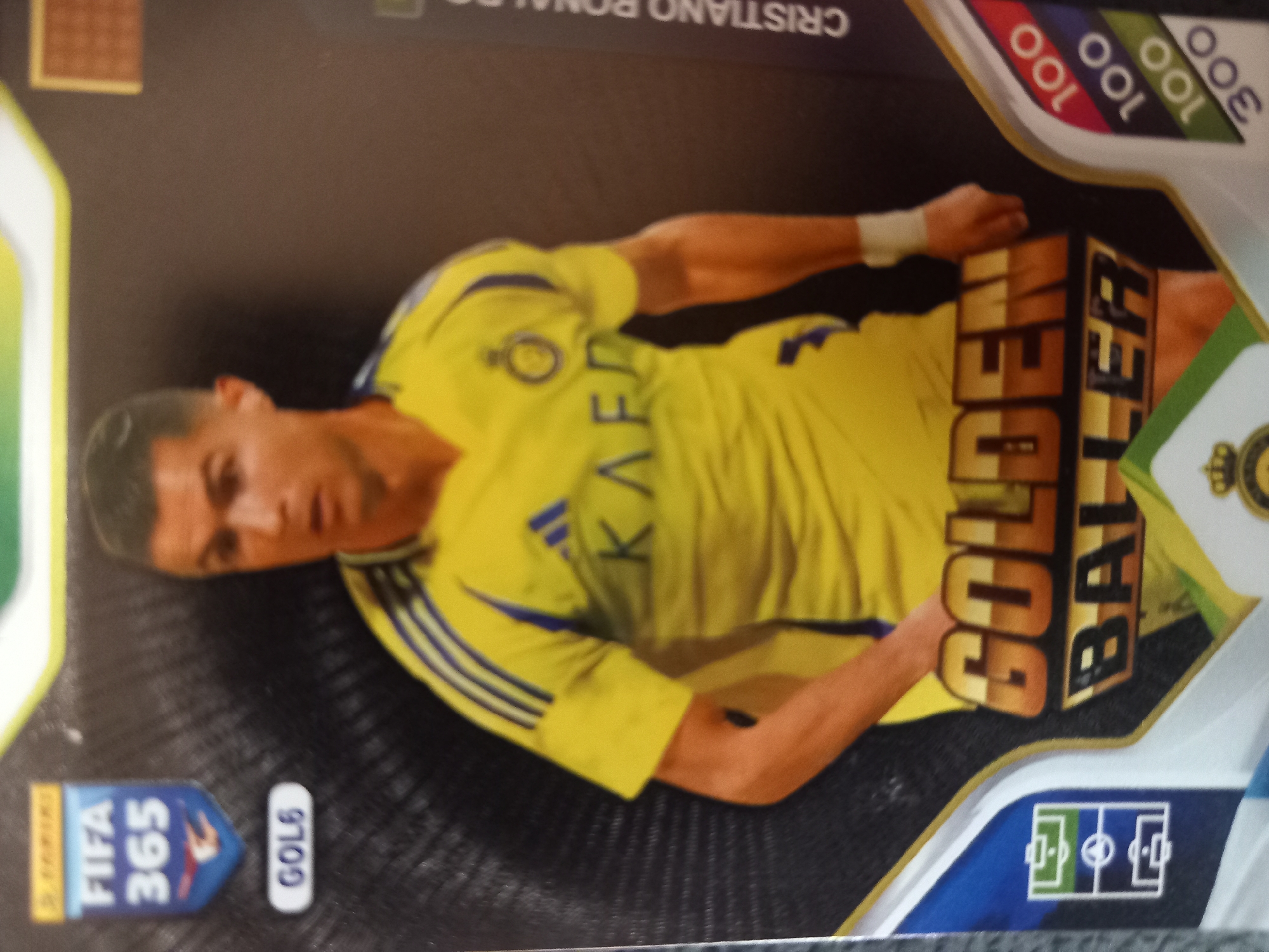 The image is a close-up, eye-level, indoor, studio shot of a trading card featuring a soccer player, Cristiano Ronaldo. The card has a black, textured background with a metallic sheen. In the upper left corner, there is a blue shield with "FIFA 365" written in white text, and a small red and white logo. Below this, a white oval shape contains the text "GOL6". Cristiano Ronaldo is wearing a yellow soccer jersey with blue stripes on the sleeves and a crest on the chest. He is looking slightly to his left, with his arms visible. Across the bottom of the card, large, golden text reads "GOLDEN BALLER". To the right of Ronaldo, vertical white text reads "CRISTIANO RONALDO". On the far right edge of the card, a series of colored bars with numbers are visible: red with "100", blue with "100", green with "100", and a darker green with "300". A stylized soccer field icon is present on the lower left side of the card.