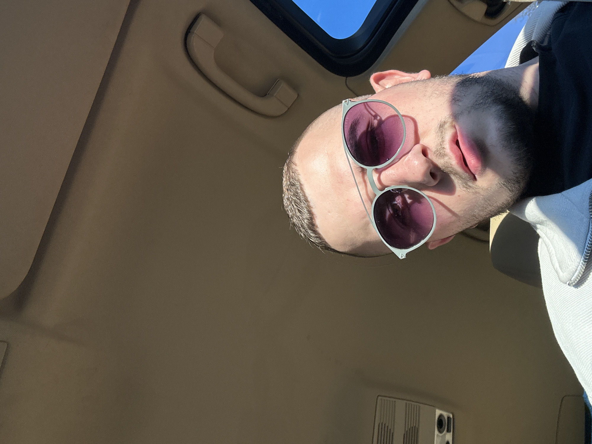A man is seen from a low angle, likely taken from the interior of a car looking up through the open sunroof. The man has short hair, a beard, and is wearing sunglasses with pink lenses. He's wearing dark clothing and possibly a light-colored jacket with a zipper visible. The interior of the car is light brown. The sky is visible through the sunroof. The location in Plovdiv, Bulgaria is not possible to determine from the provided image.