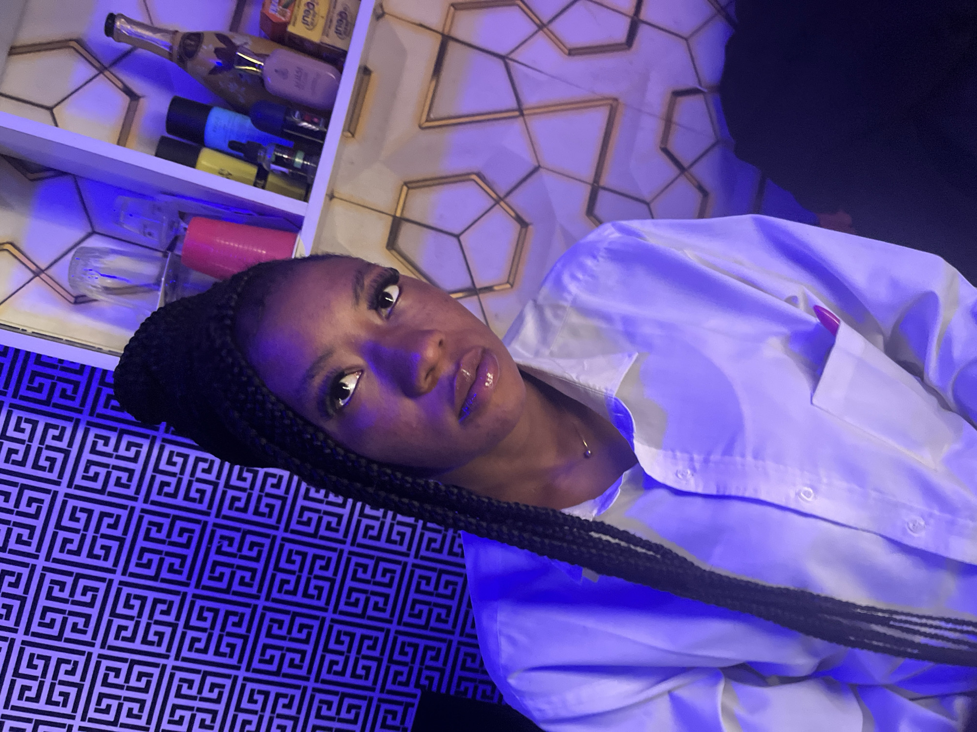 The media file shows a close-up, high-angle, portrait-oriented shot of a young Black woman. She is looking directly at the camera with a neutral expression. Her hair is styled in long, dark braids, some of which are draped over her shoulder and across her chest. She is wearing a white button-down shirt, open at the collar, revealing a delicate gold necklace. Her lips are glossy and appear to be subtly colored.
The background is a mix of elements. To the left, a white shelf unit is visible, containing various bottles and objects, some lit by blue and purple lights, suggesting an indoor, possibly nightlife or retail setting. One bottle appears to be a champagne bottle. The wall behind the shelf has a geometric pattern with gold accents. To the right of the woman, a dark, blurry area suggests more of the environment, possibly a shadowed corner or furniture. The lower portion of the image is dominated by a black and white patterned surface with a repeating geometric design, illuminated by blue light. This patterned surface could be wallpaper, a textured wall, or fabric.
The lighting is dramatic and moody, with strong blues and purples creating an atmospheric effect. The time of day is not explicitly indicated, but the artificial lighting suggests it could be evening or night. There is no visible text in the media file. The overall impression is one of a stylish and perhaps intimate setting.