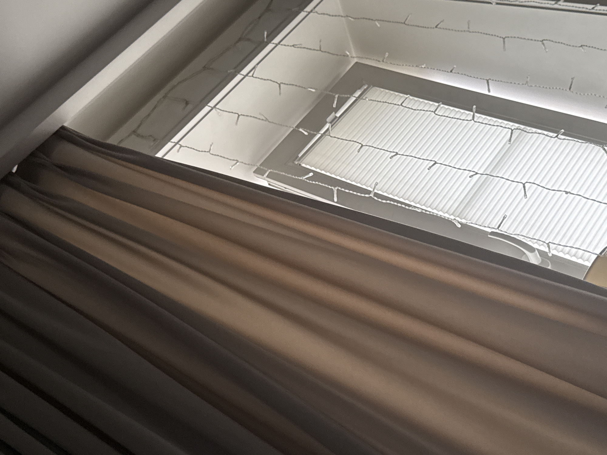 The image shows a portion of a room with a window. On the left side of the frame, a brown curtain hangs diagonally, obscuring a large part of the scene. Through the gap in the curtain, a window with white horizontal blinds is visible. Along the top of the window and extending downwards, a string of small, white lights is draped. The lighting suggests it is daytime, with light coming from the window. The overall environment appears to be indoors, possibly a bedroom or living area. The location is unknown.