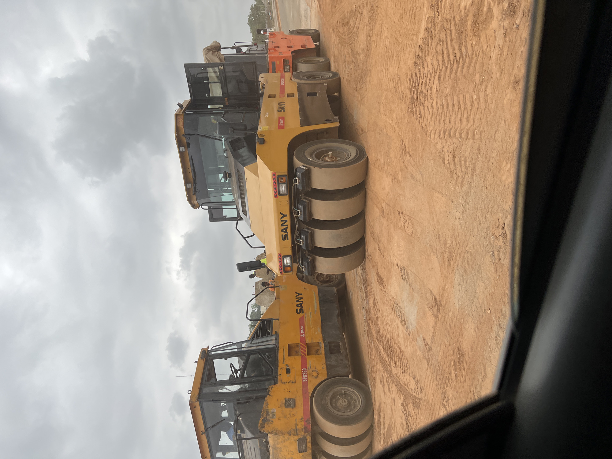 This is a daytime photograph captured at a construction site in Abuja, Nigeria, under an overcast sky. The image primarily features a lineup of heavy construction machinery, specifically road rollers, on a reddish-brown dirt surface.
In the foreground and middle ground are two large, yellow SANY road rollers (also known as pneumatic tire rollers or soil compactors). The roller closest to the viewer displays the model number "SPR260" on its side, while the one behind it has "SMH" visible, likely part of its model designation. Both rollers feature multiple large, smooth pneumatic tires designed for soil compaction. A person is dimly visible inside the cab of the foreground roller, and another person's head can be seen near the cab of the middle roller.
Further in the background, a smaller orange construction vehicle is partially visible. The ground is a dry, dusty reddish-brown earth, showing distinct patterns from the rollers' tires and other vehicle tracks, indicating ongoing compaction work. The sky overhead is uniformly gray and cloudy, suggesting an overcast day. The overall scene depicts a typical heavy civil engineering construction environment.