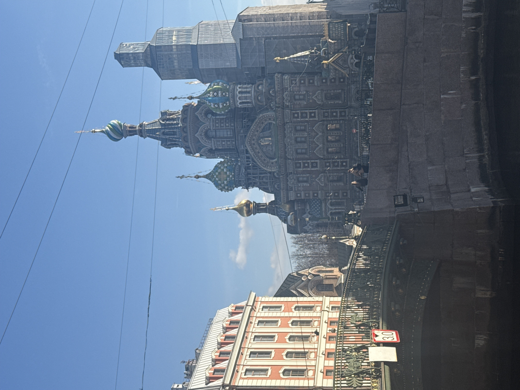 The Church of the Savior on Spilled Blood in Saint Petersburg, Russia, stands prominently beside a canal. Scaffolding surrounds a portion of the church. A bridge with ornate metalwork spans the canal, displaying a speed limit sign. A building with red and white facade is visible below the bridge. Wires stretch against a blue sky. Two people are visible on the embankment.