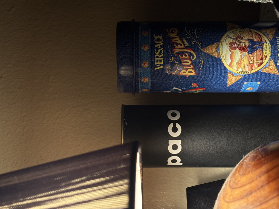 The image depicts a dimly lit indoor scene featuring three primary objects against a warm-toned, textured wall. On the far left, the lower portion of a pleated lampshade, illuminated from within, is visible, casting light towards the center. In the immediate lower-left foreground, a blurred, curved object with a light brown, wood-grain texture is partially visible. Centered in the image is a tall, black cylindrical container with the white sans-serif text "paco" prominently displayed vertically on its side. To the right of this, another tall, blue cylindrical container stands. This container features a denim-like pattern and is adorned with various decorative elements. Prominent text on the blue container includes "VERSACE" at the top, followed by "Blue Jeans MAN" in stylized lettering. Further down, a star-shaped motif frames an illustration of a cowboy playing a guitar in a desert landscape. Below this illustration, "EAU DE TOILETTE" is visible. There are no discernible visual cues within the image to specifically identify the location as Tanger, Morocco.
