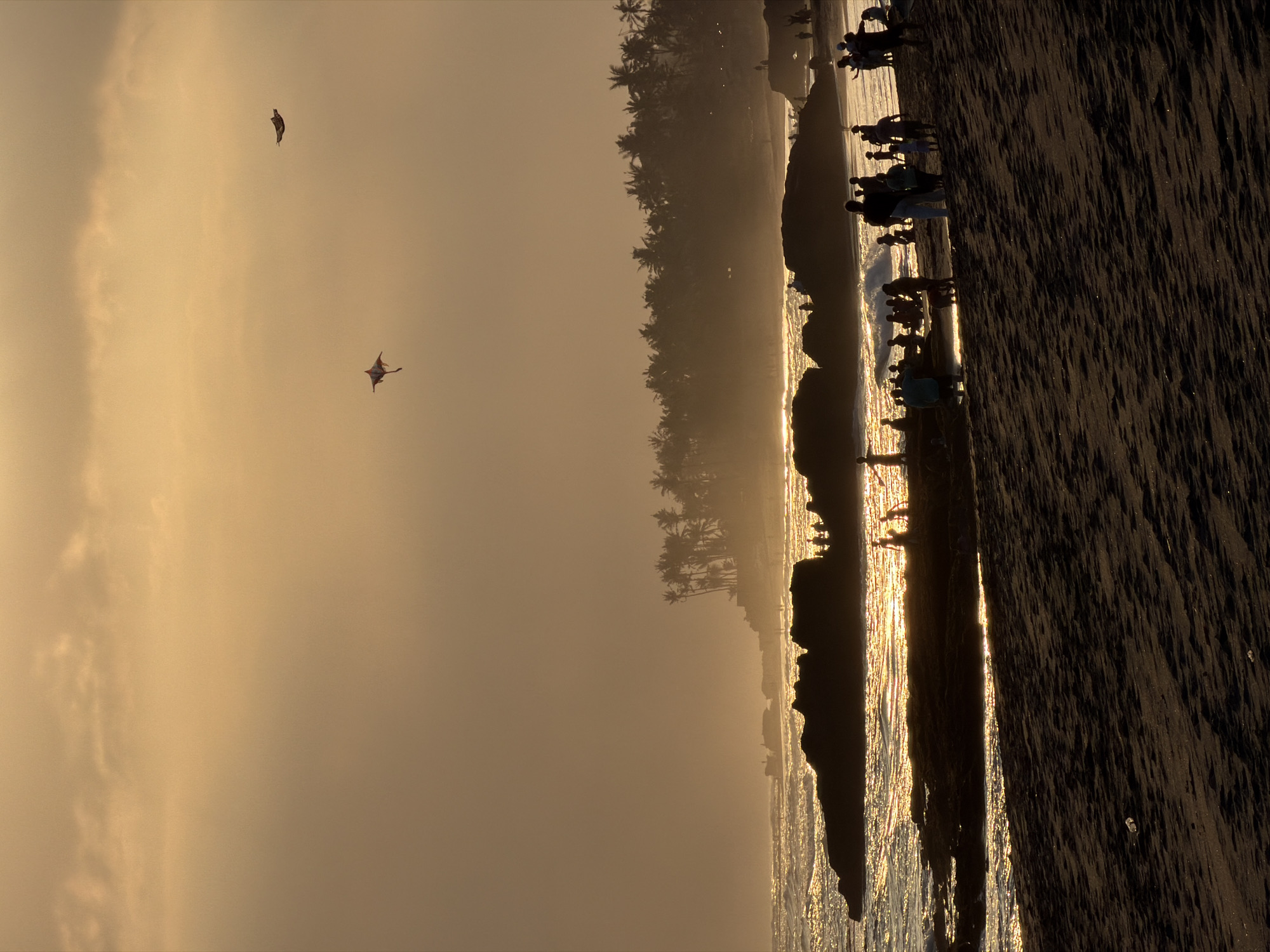 The image depicts a silhouetted scene of people on a beach in Mengwi, Indonesia, during sunset. The sky is a warm, hazy orange, with the sun casting a golden glow on the ocean waves and wet sand. Several palm trees line the distant shore, silhouetted against the sky. In the foreground, people are gathered on the dark sand, some walking, others standing in groups. The ocean has light waves crashing against dark rocks in the middle ground, where more people are visible. Two kites are flying in the sky; one is a diamond-shaped kite with a white and red pattern, and the other is a darker, wing-shaped kite. The overall atmosphere is peaceful and serene, capturing a moment of leisure at the end of the day.