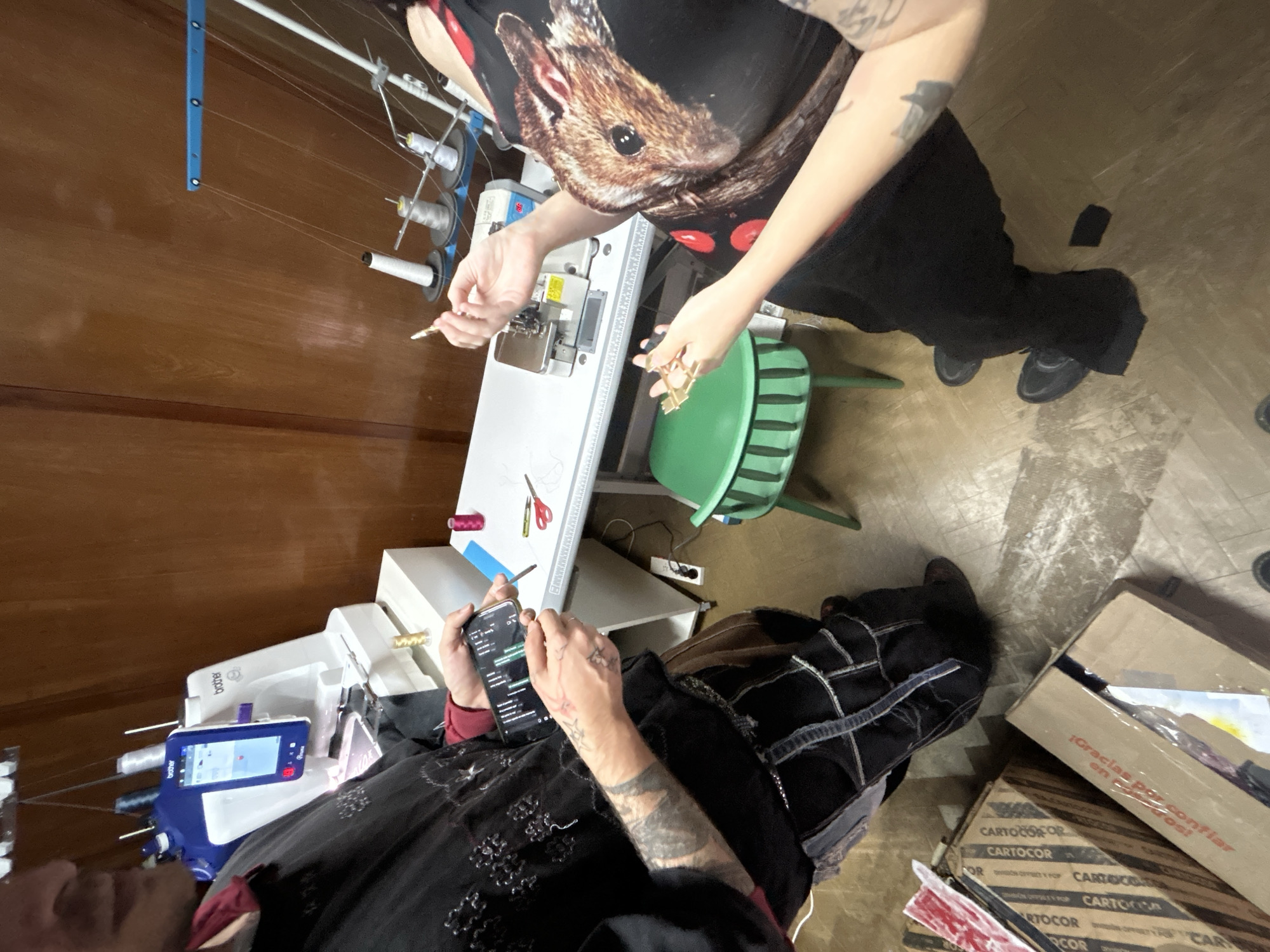 The scene depicts two individuals in what appears to be a sewing or crafting space. The image is taken from an overhead perspective.
One person is wearing a dark-colored top with a print of a rodent on it and black pants. They are standing at a sewing machine with a white surface, and they appear to be manipulating small golden objects.
The second person is seated and wearing a black outfit. They are holding a smartphone in their hands, presumably interacting with it.
Several sewing machines, spools of thread, scissors, and other crafting tools are visible. The background includes wooden paneling on the wall and flooring with a mottled pattern. A green chair is also present.
In the lower-right corner, there are cardboard boxes stacked on top of each other, one bearing the inscription "¡Gracias por confiar en Carbos!". The inscription suggests the location is in a Spanish-speaking region, potentially Argentina.