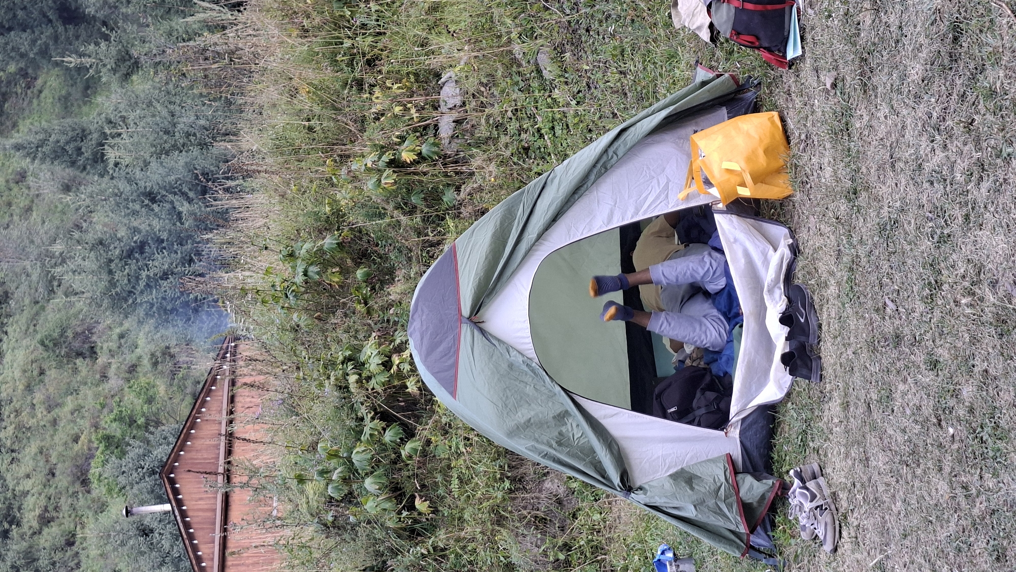 A person's legs and feet, wearing blue and orange socks and light-colored pants, are visible protruding from the entrance of a green and white tent pitched on a grassy, uneven surface. The tent is set up in a natural environment, with a dense forest and vegetation covering a hillside in the background. To the left of the tent, a pair of gray athletic shoes rests on the grass, and a blue object is partially visible. Inside the tent, dark bags and bedding are present. To the right of the tent, a bright yellow tote bag and a backpack with red straps are placed on the ground. In the upper left portion of the image, a wooden structure resembling a cabin with a chimney is visible amidst the trees and vegetation, with faint smoke emanating from the chimney. The overall scene depicts a campsite in a remote, possibly mountainous or forested area. The location is not specified to be in the city of Batan, Kazakhstan.