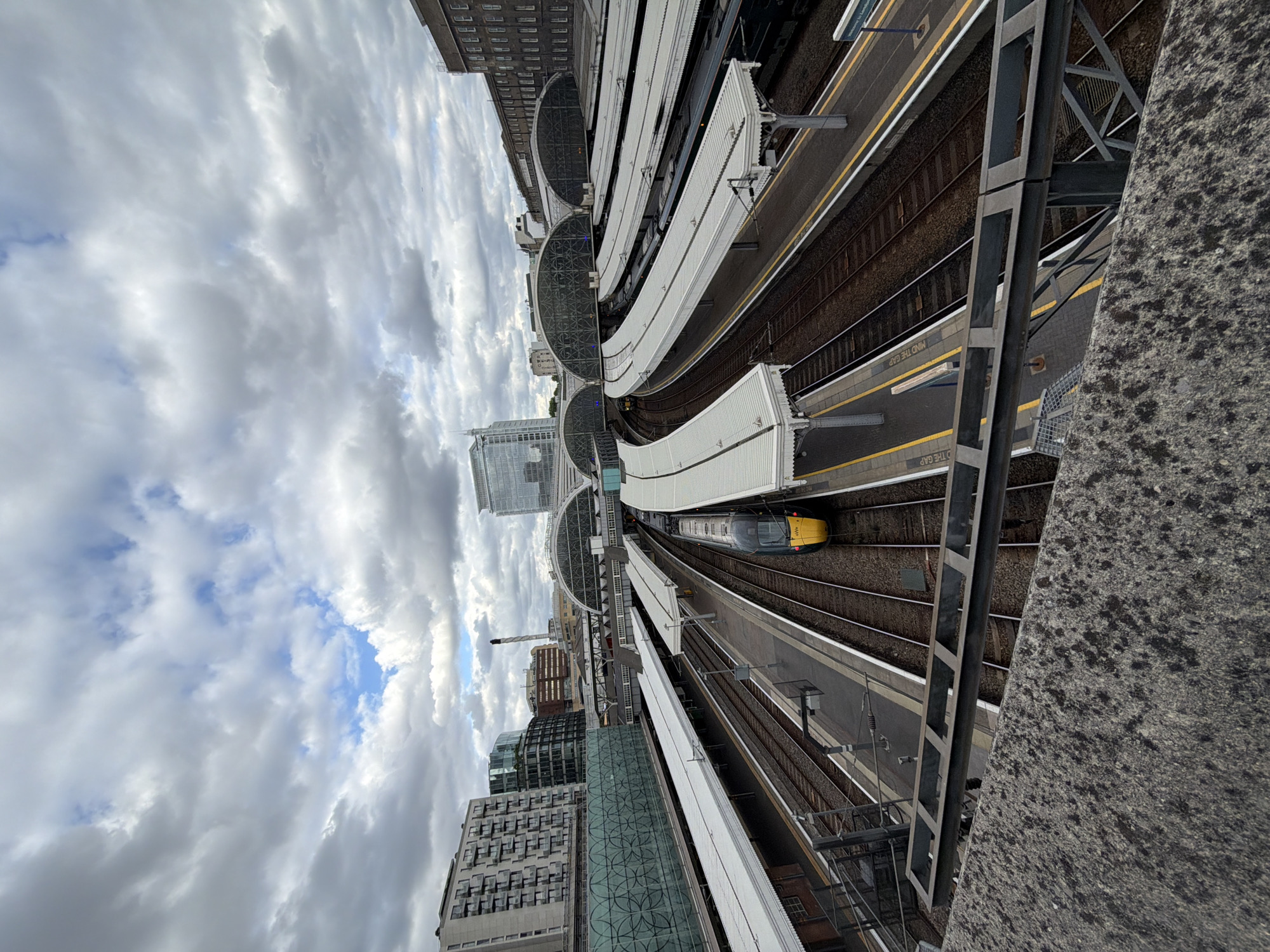 This is an elevated view of Paddington Station in London, United Kingdom. The shot is taken from a high vantage point, possibly from a bridge or a tall building overlooking the station. The foreground shows a concrete barrier or wall. Numerous train tracks and platforms can be seen, with white canopies covering parts of the platforms. A yellow and gray train is visible on one of the tracks. The sky is filled with clouds, with patches of blue visible. Several buildings of varying sizes and architectural styles surround the station, indicating the urban setting. The shot is taken in daylight.