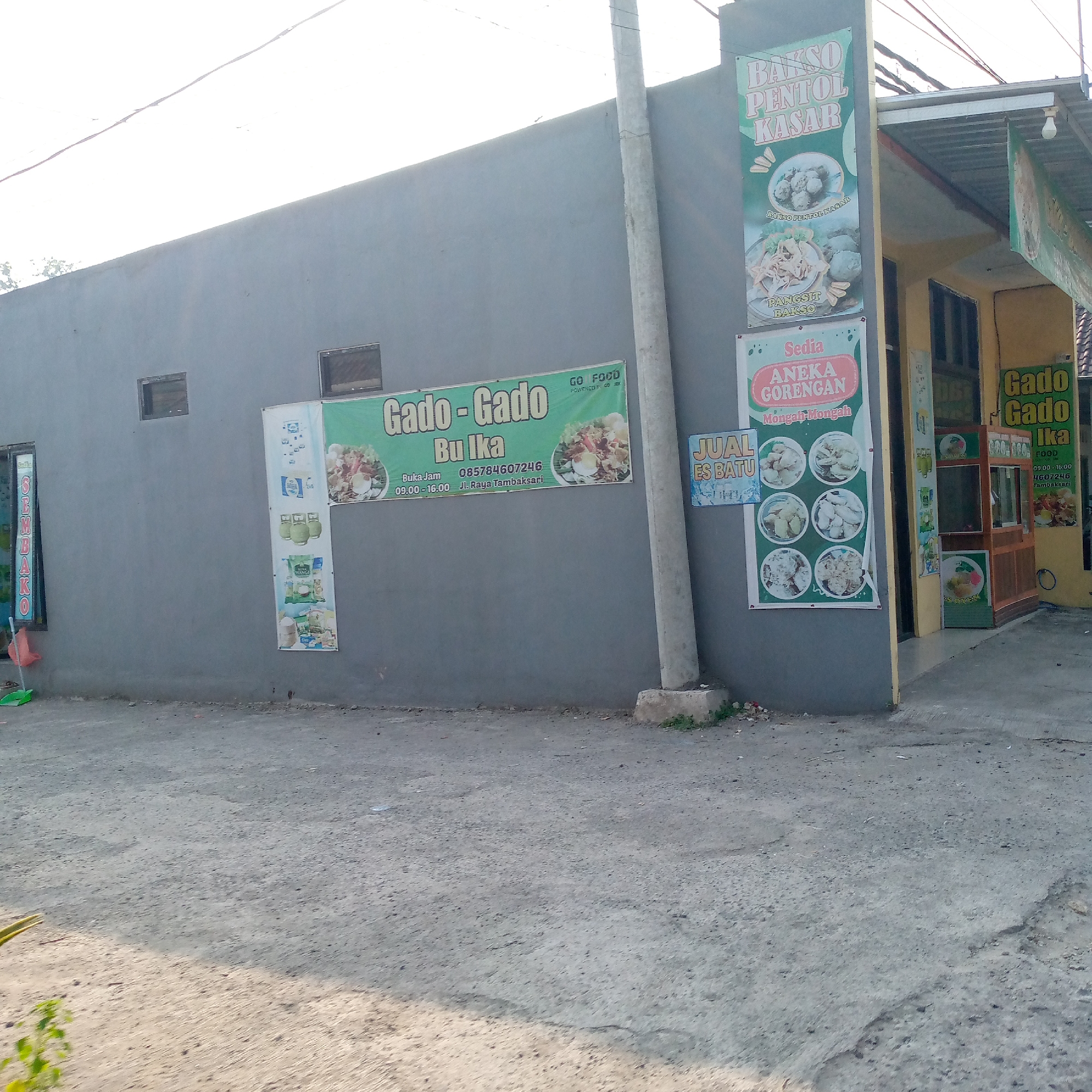 The image shows a grey building with various advertisements for food items. On the left side of the building, a vertical sign reads "Sembako" which typically means groceries. Further to the right, a large green banner advertises "Gado - Gado Bu Ika" with contact information and operating hours from 09:00 to 16:00. Below this banner, another vertical sign on a lamppost advertises "BAKSO PENTOL KASAR" and "PANGSIT BAKSO". Next to that, another green banner advertises "Sedia ANEKA GORENGAN" (Fried Snacks) with pictures of various fried foods and a "JUAL ES BATU" (Ice Cubes for Sale) sign. To the far right, another "Gado Gado Ika" sign is visible. The scene appears to be outdoors, with a paved area in the foreground. The building itself has a yellowish awning extending from the right side. The location is identified as Tambakagung, Indonesia. There are no people visible in the image.