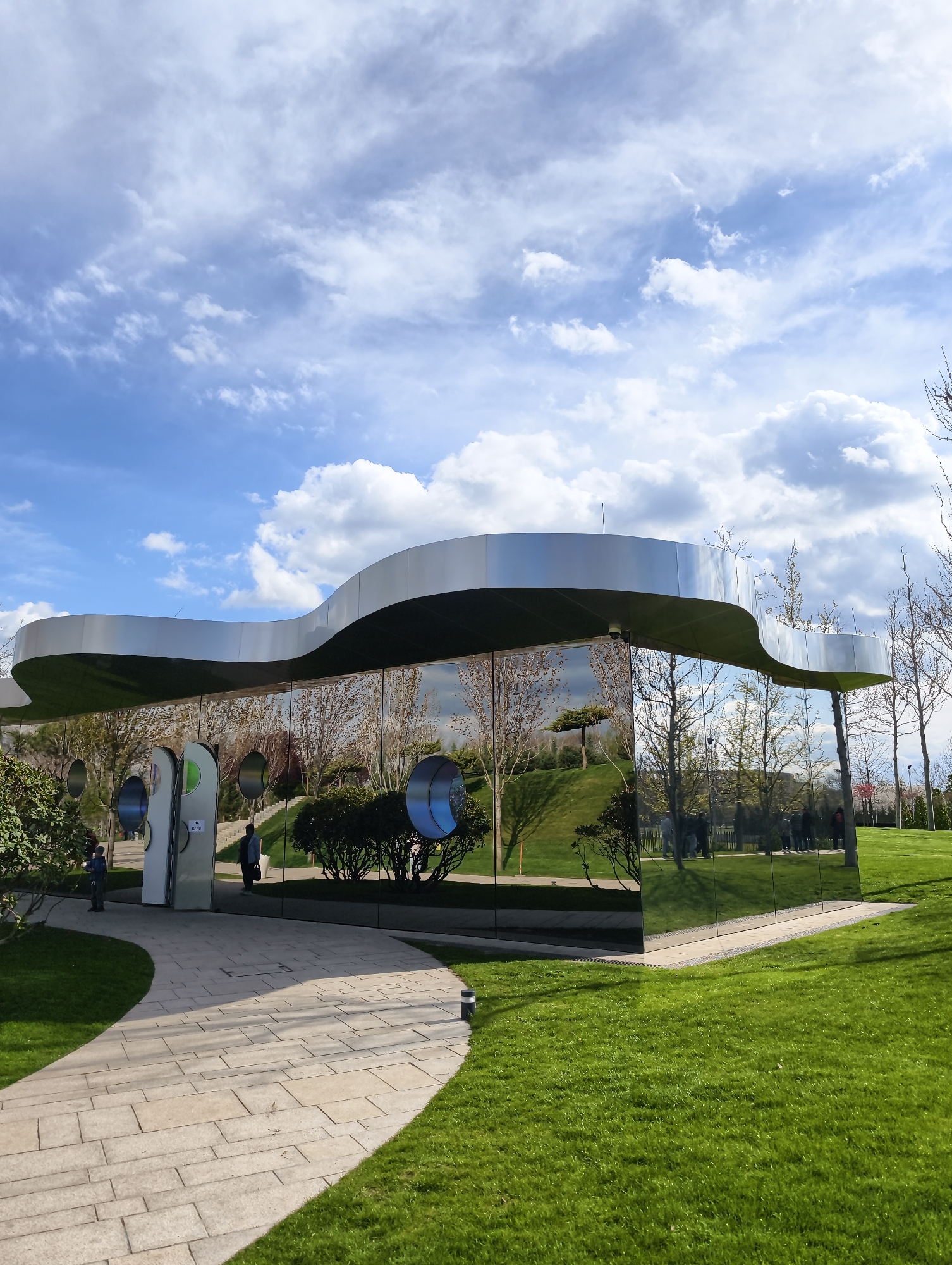The image captures a modern, distinctively designed building situated within a vibrant, well-maintained park under a partly cloudy blue sky. The building features a sinuous, wave-like metallic roof structure, possibly made of silver-colored metal. Its walls are composed entirely of highly reflective glass, mirroring the surrounding landscape, including the sky, fluffy white and gray clouds, lush green grass, and bare trees, creating a visually striking integration with nature.
Several circular, metallic elements are embedded into the reflective facade, some with colorful inner rings (green, blue). A light-colored paved pathway, made of rectangular stones, curves gently towards the building's entrance from the left foreground. To the right and around the building, well-manicured green grass extends, sloping gently into the background.
A few individuals are visible, either directly or as reflections in the glass. One person walks along the paved path towards the building, while another stands near the entrance on the left. In the reflections, more people can be seen in the park's background. The scene depicts a calm daytime setting, likely in spring given the mix of bare and budding trees.
A small white sign near the entrance is visible, with the text "НА СЕБЯ", which translates to "TO MYSELF" or "PULL", indicating how to open the door. This architectural structure is located in Krasnodar, Russia, suggesting a contemporary public space or facility.