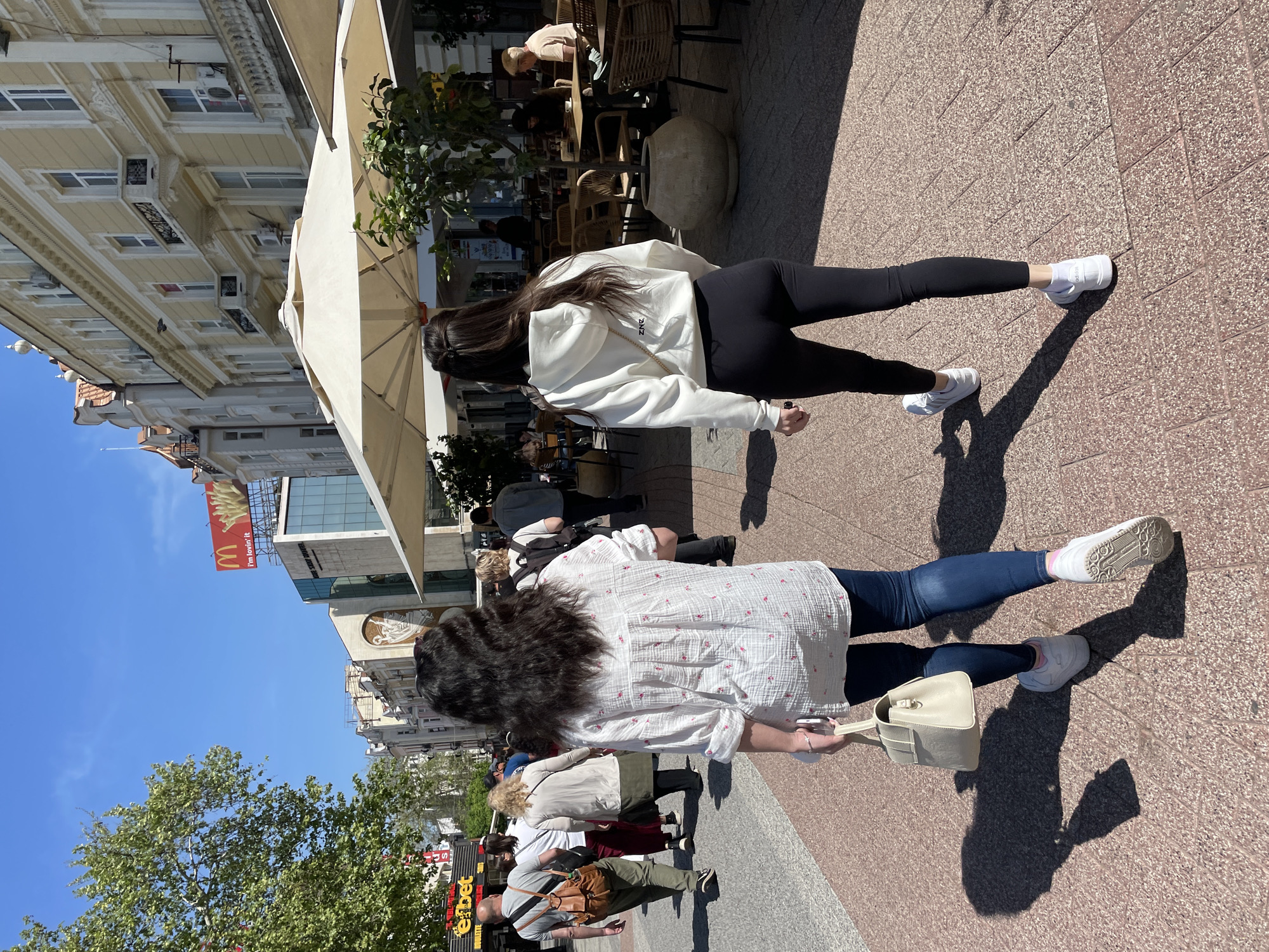 This image captures a vibrant, sunny day in a pedestrian area of Plovdiv, Bulgaria, featuring two women walking in the foreground, viewed from behind. The scene suggests a bustling urban environment with outdoor cafes and historic architecture.
The woman closer to the viewer has dark, curly hair and wears a white, patterned button-up shirt with small pink or red designs, blue jeans, and white sneakers. She carries a light beige handbag on her right arm and appears to be holding a phone in her left hand. Her white sneakers have visible textured soles.
Just ahead of her, the second woman has long, straight dark hair and is dressed in a white hooded sweatshirt, black leggings, and white sneakers. A thin strap, possibly from a small shoulder bag, is visible over her right shoulder. Text, possibly "2NZ," is subtly visible on the back of her hoodie. Both women cast long, distinct shadows on the light brown paved ground, indicating bright sunshine, likely during the mid-morning or afternoon.
To the left, an outdoor cafe or restaurant with wicker chairs and tables is shaded by a large, light-colored umbrella. A large ceramic pot sits near the cafe seating. Beyond the cafe, multi-story buildings with numerous windows, balconies, and air conditioning units line the street, showcasing European architectural styles. In the distance, a large red McDonald's billboard with the golden arches and "I'm lovin' it" text is visible against a clear blue sky. Further down the street, an "efbet" sign can be seen, hinting at local businesses. Green trees add to the urban landscape. Other pedestrians are visible further back, contributing to the lively atmosphere.