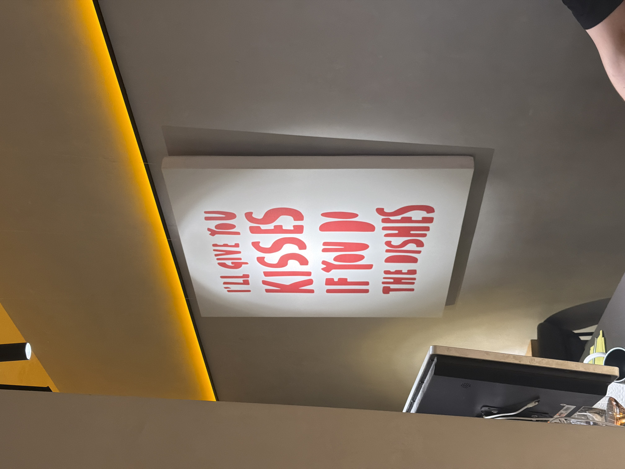 The image, taken in Krasnoyarsk, Russia, displays an indoor scene, likely a contemporary kitchen or dining area in a cafe or home. The central focus is a white rectangular sign with bold red capital letters, which reads: "I'LL GIVE YOU KISSES IF YOU DO THE DISHES".
The sign is mounted on a light grey wall or ceiling. To its left, a bright, warm yellow LED strip illuminates an architectural edge, casting a soft glow. In the lower right corner, a section of a light-colored counter or shelf is visible, upon which rests a dark-colored monitor or tablet with several white cables connected to its rear. A person's bare arm and shoulder, partially visible in the upper right, wearing a black sleeve, suggest an individual is present nearby. The overall ambiance is clean and modern, with no specific activity captured, but the sign hints at domestic or communal interaction.