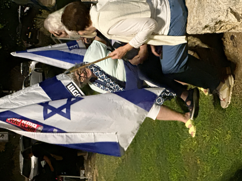 The image displays an outdoor nighttime scene in Mishmar Hasharon, Israel, featuring three elderly women gathered on a grassy area. In the center-left, an elderly woman with grey hair and glasses, wearing a light-colored top, dark pants, a blue and white patterned scarf, and yellow sandals, smiles while looking to her right. She is partially obscured by a large Israeli flag, which bears a red sticker with white Hebrew text "עם ישראל חי" (Am Yisrael Chai) and blue Hebrew text "חופש" (Chofesh - Freedom) below its blue Star of David. To her right, an elderly woman with light-colored hair, dressed in a white long-sleeved top, a white knitted vest, and blue pants, sits on a light-colored stone ledge. She holds a slender wooden stick in her right hand and a mobile phone is visible near her left thigh. A third elderly woman with grey hair is partially visible behind the second woman, engaging in conversation. Another Israeli flag, also featuring the Hebrew word "חופש" (Chofesh - Freedom), stands in the mid-background to the right of the first woman. The foreground is a grassy lawn illuminated by artificial light. In the dark background, several vehicles are present, with a person in dark clothing partially visible bending near a dark car on the far left.