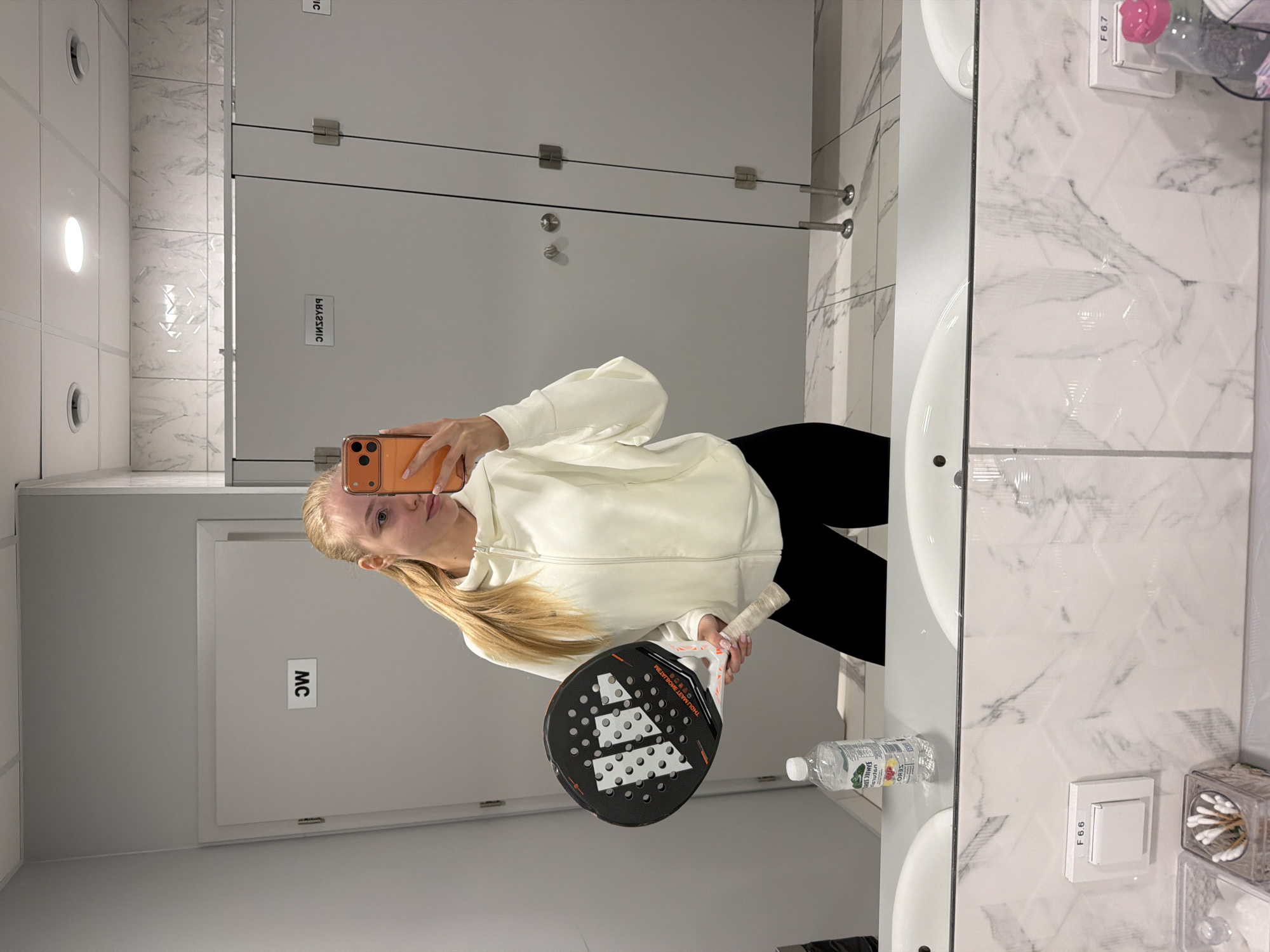A woman is taking a selfie in a public restroom. She is holding a paddle tennis racket in her left hand. The bathroom has tiled walls, and there is a row of stalls in the background. There is a mirror in front of the woman. A bottle of water is visible next to the sink.